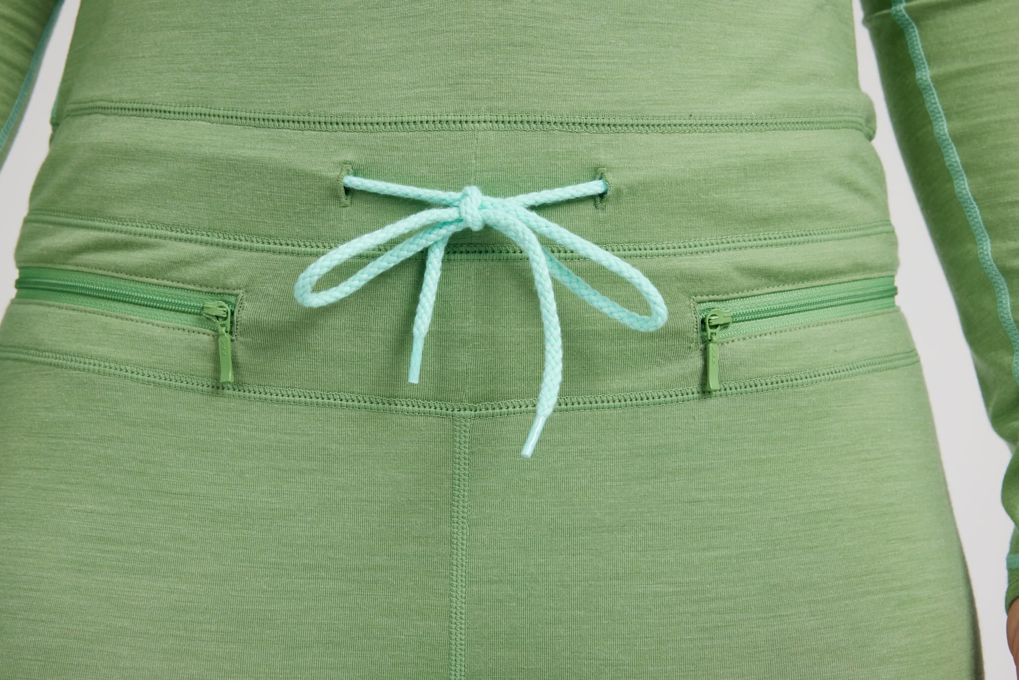 Close-up of the back waist of olive green athletic pants featuring two zippered pockets and a mint green drawstring tied in a bow.
