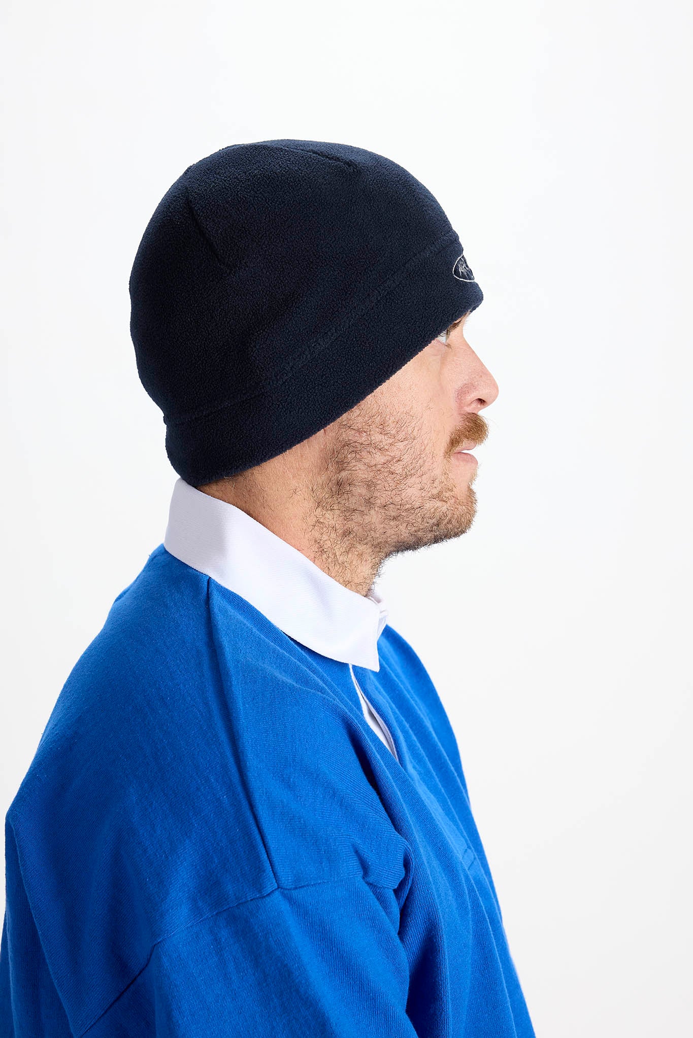 A man in profile wearing a black knit beanie, a blue long-sleeve shirt, and a white collared shirt, photographed against a plain white background.