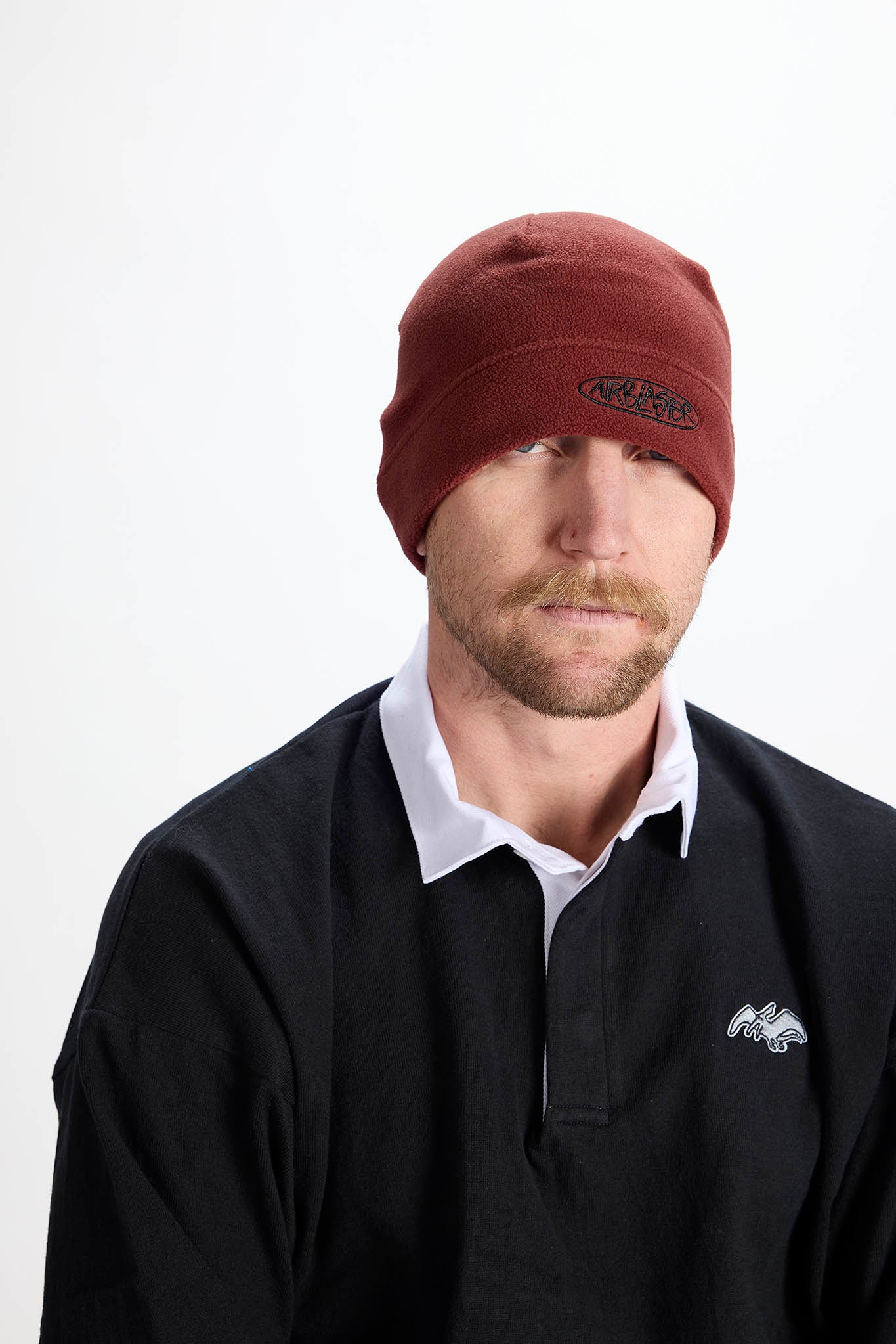 A man wearing a dark red knit beanie with a small embroidered logo on the front, paired with a black long-sleeve rugby shirt over a white collared shirt, photographed against a plain white studio background.