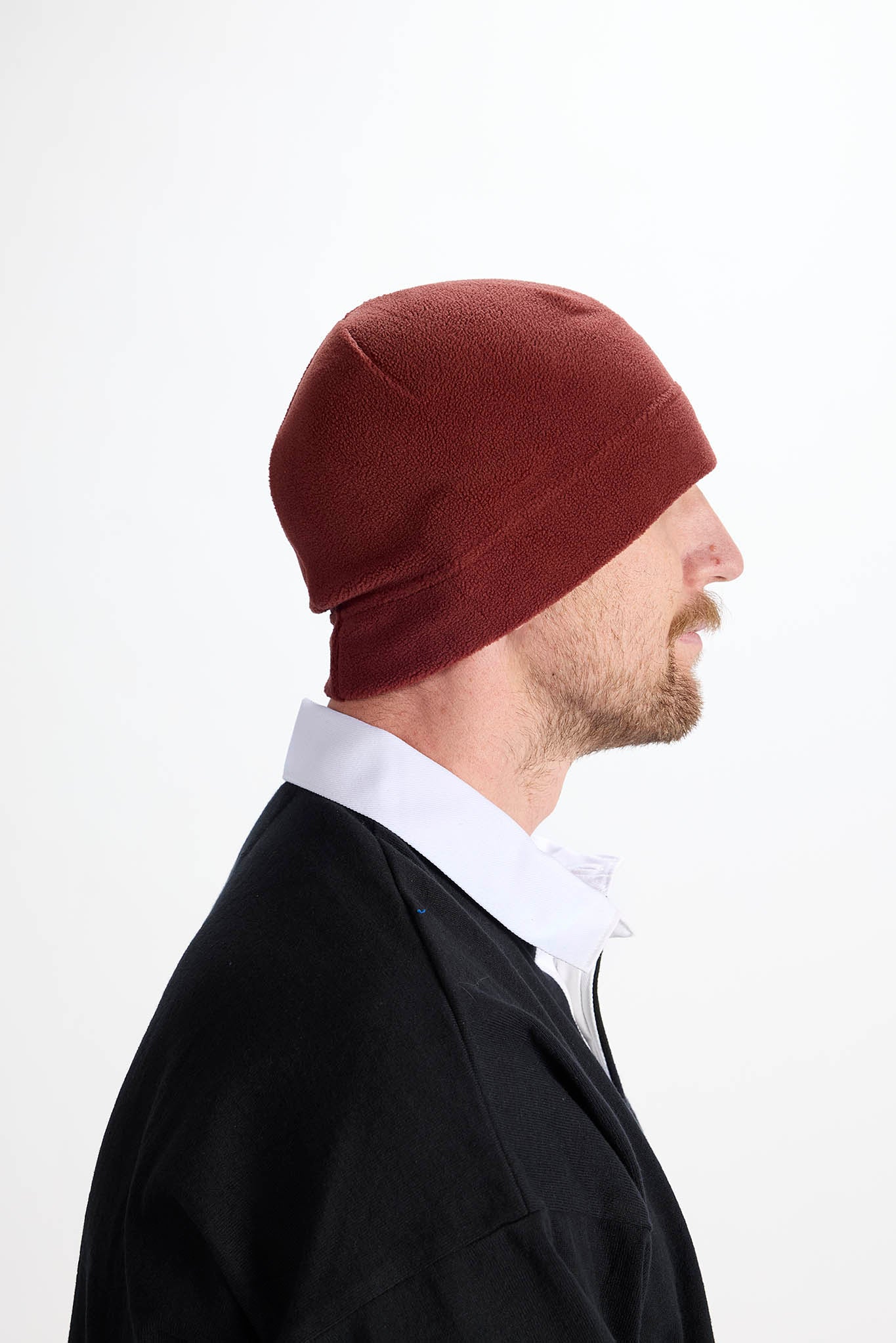 A man in profile wearing a dark reddish-brown knit beanie, a white collared shirt, and a black jacket, photographed against a plain light gray background.