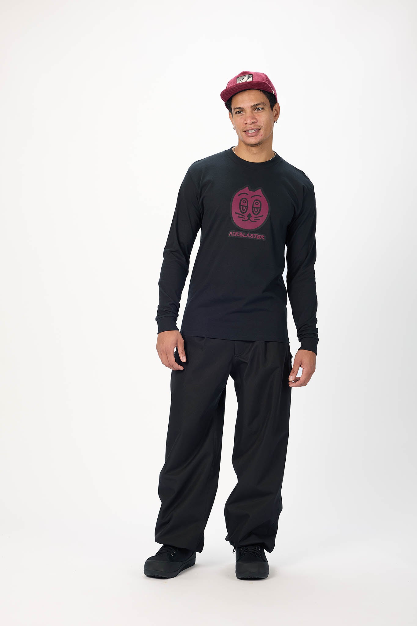 A man modeling a black long-sleeve t-shirt with a pink circular graphic on the chest, black pants, and a maroon baseball cap, standing against a plain white studio background.