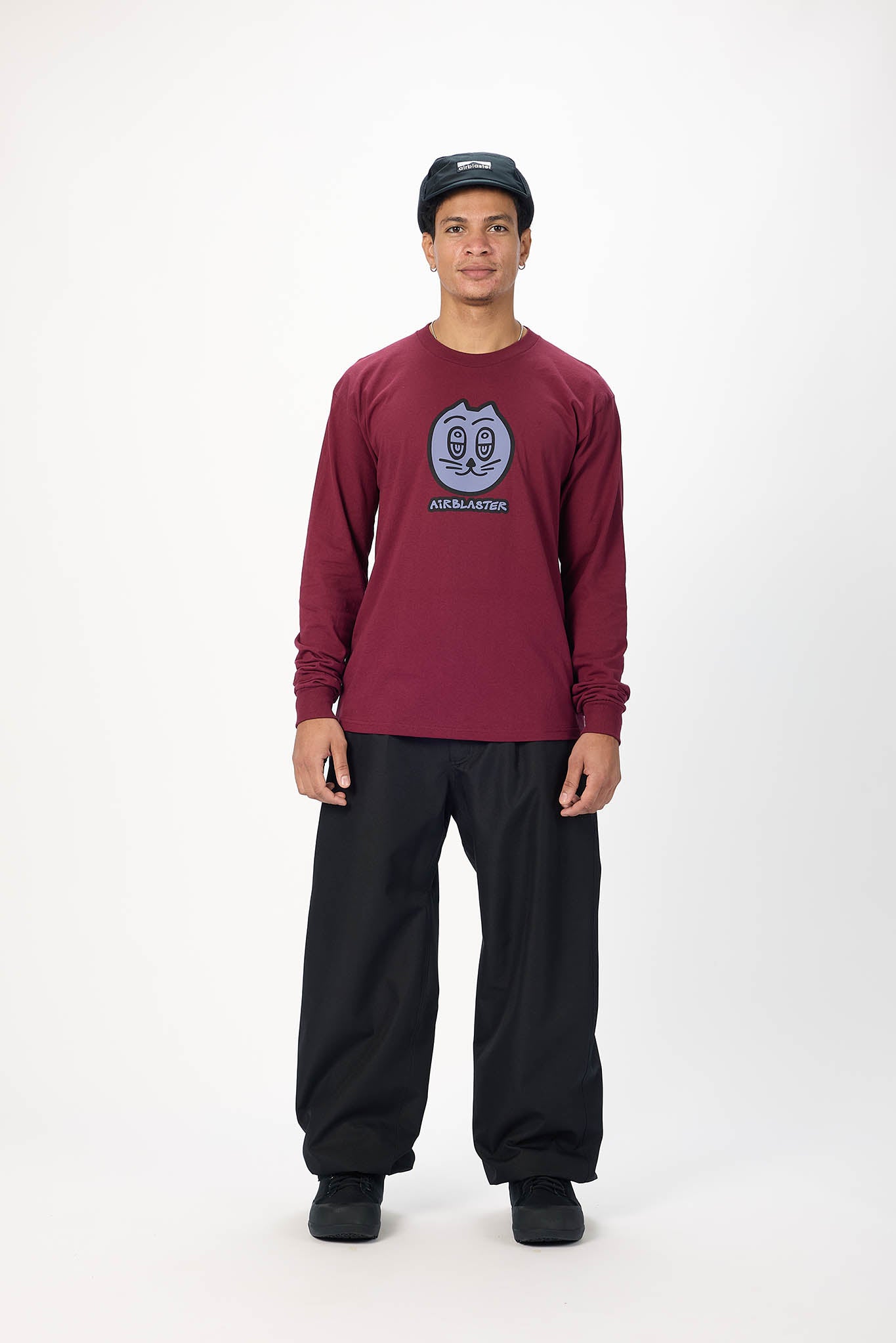 A man modeling a burgundy long-sleeve graphic t-shirt with a circular logo on the chest, paired with black sweatpants and a black baseball cap, against a plain white studio background.
