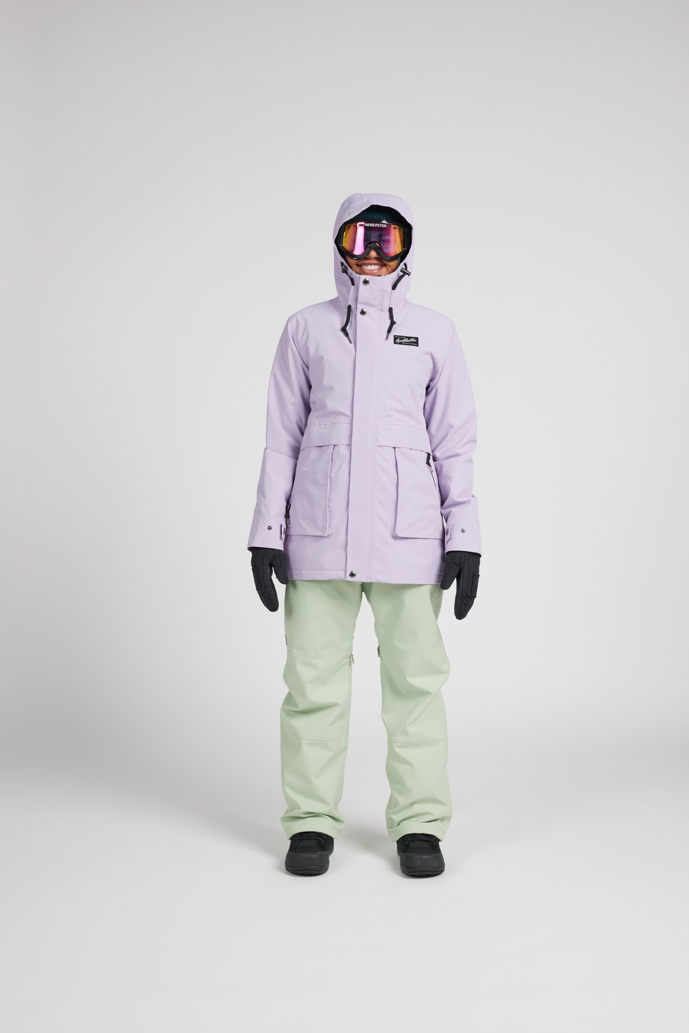 A model stands against a light gray studio background wearing a lavender hooded snowboard jacket, sage green snow pants, black gloves, and black boots. The jacket has a small black rectangular logo patch on the left chest.