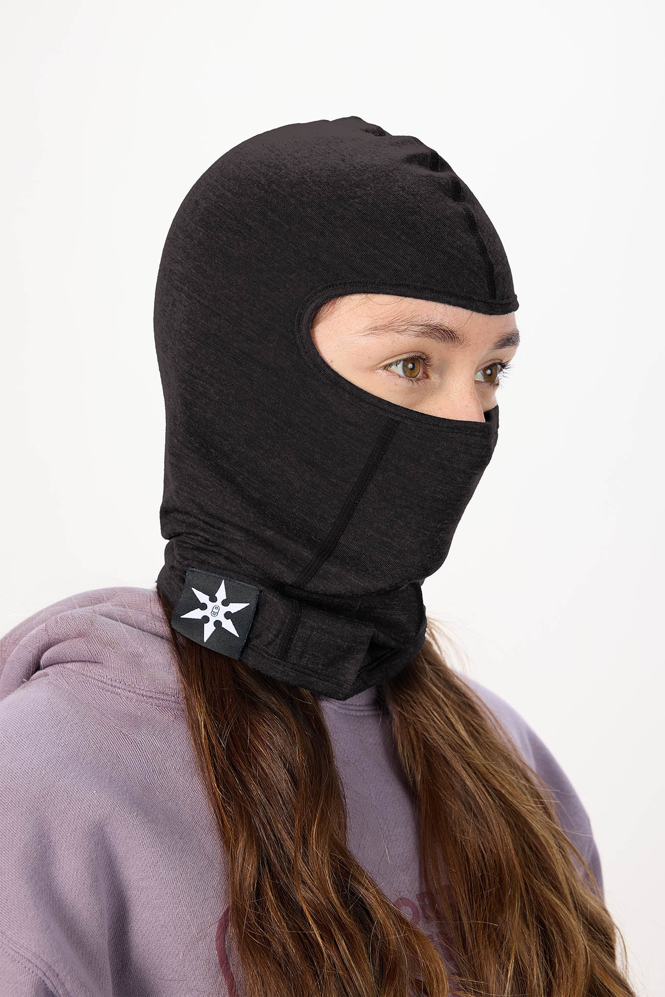 A person wearing a black full-head balaclava hood with a face opening and a lower neck flap featuring a white geometric logo, photographed against a plain light background.