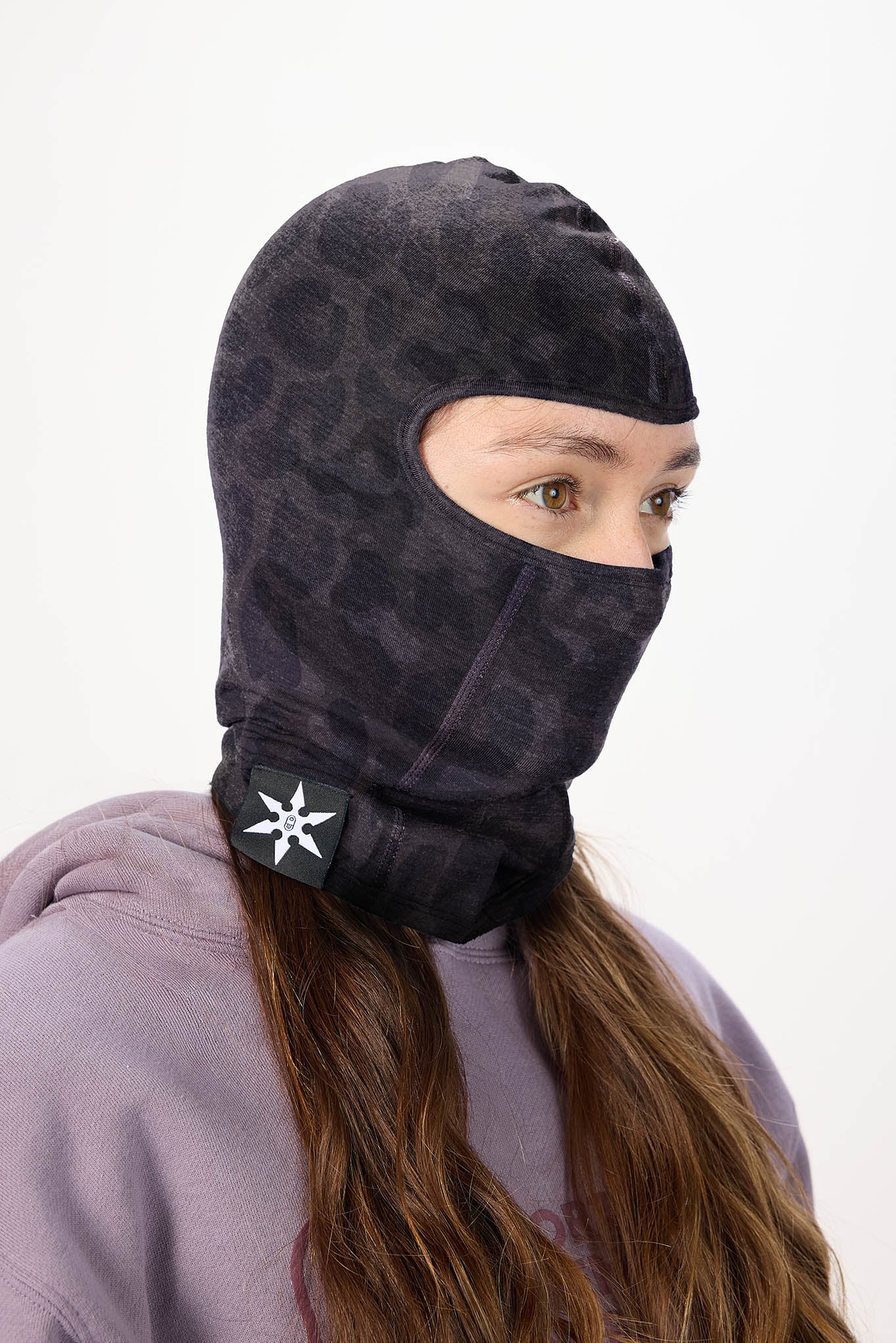 A person wearing a dark patterned full-head balaclava mask with a neck gaiter and a black logo patch, paired with a lavender hoodie against a plain white background.