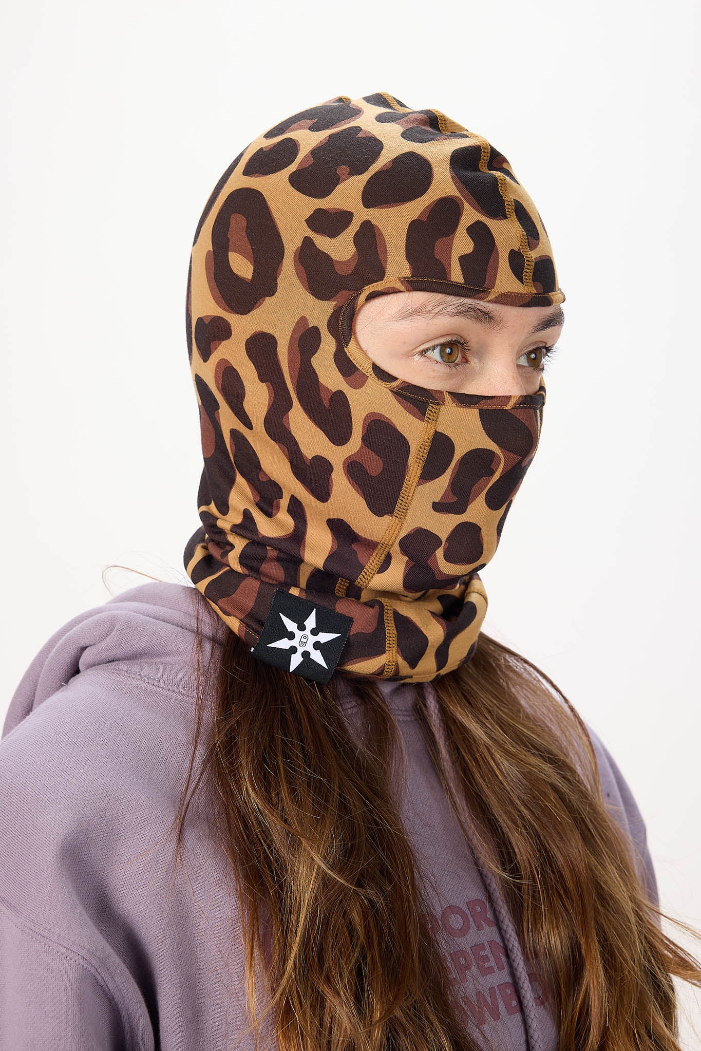 A person wearing a leopard-print balaclava hood with a cutout for the eyes and nose, paired with a lavender hoodie against a plain white background.