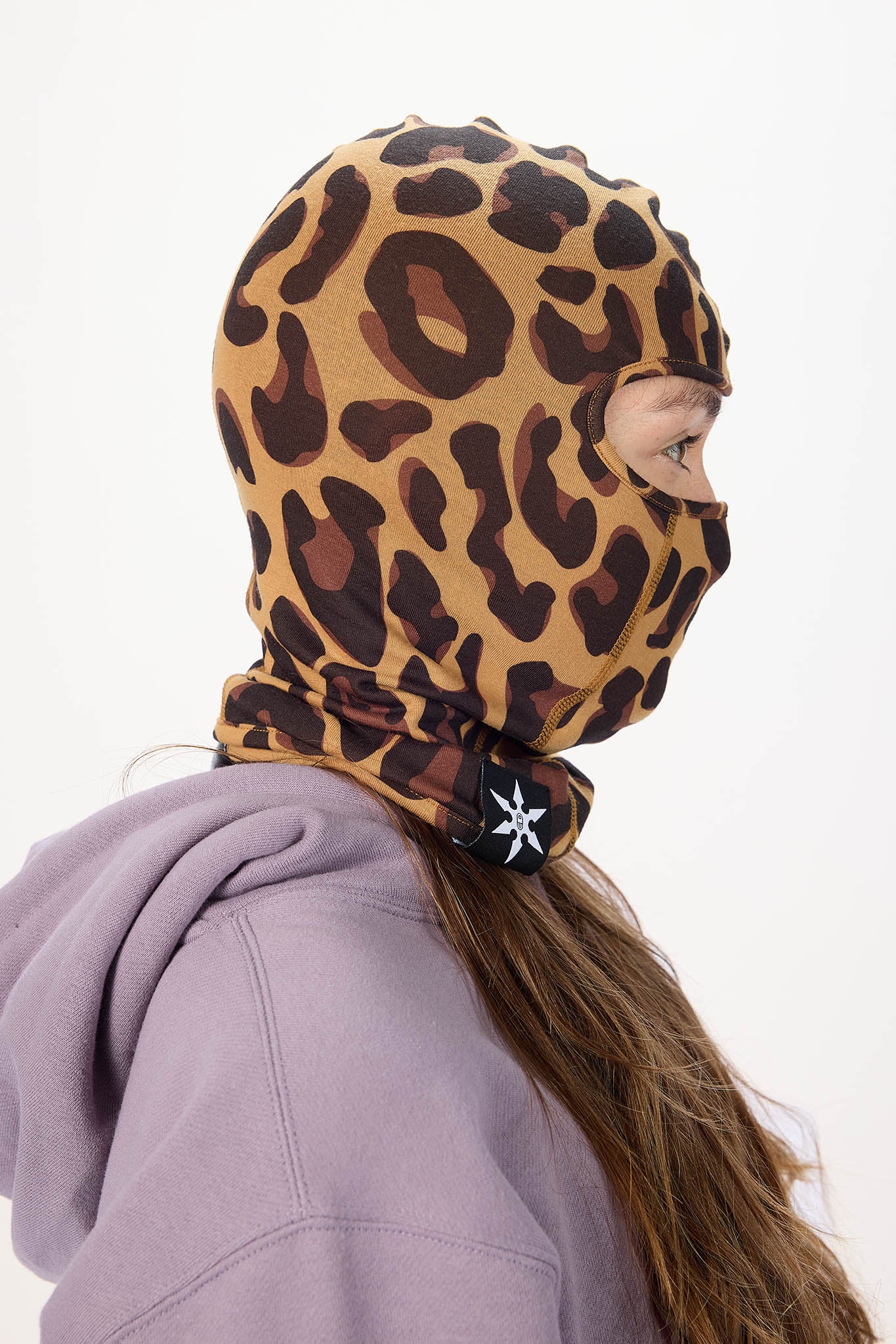 A person shown in side profile wearing a leopard-print balaclava hood and a lavender hoodie against a plain white background.