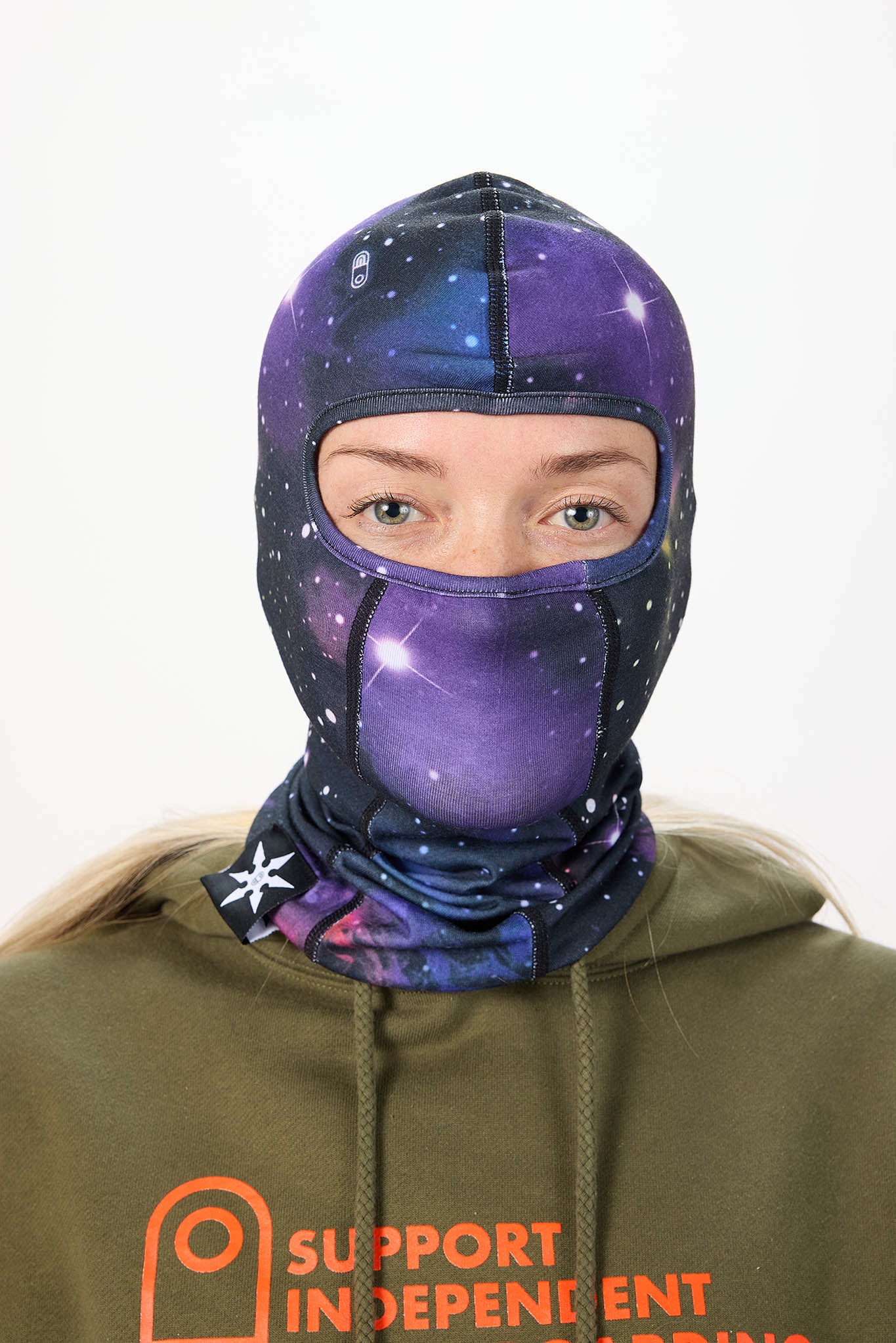 A person wearing a purple and blue galaxy-print balaclava face mask over an olive green hoodie, shown against a plain white background.