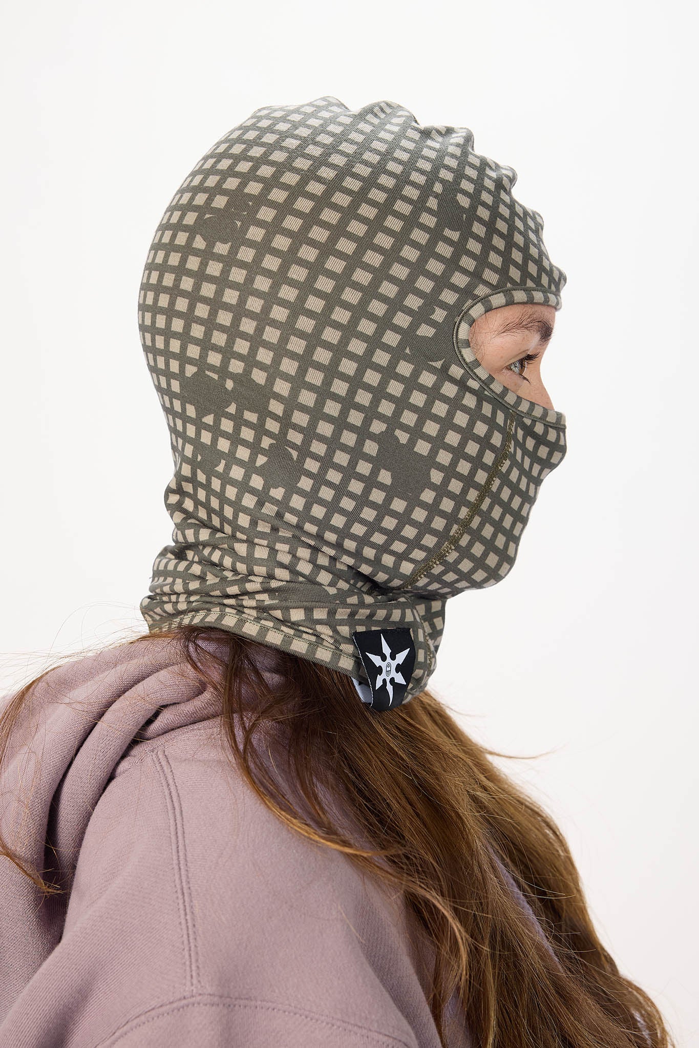 A person shown in side profile wearing a full-head patterned balaclava mask. The mask has a muted olive green and light gray grid pattern, and a small black logo patch is visible at the neck.