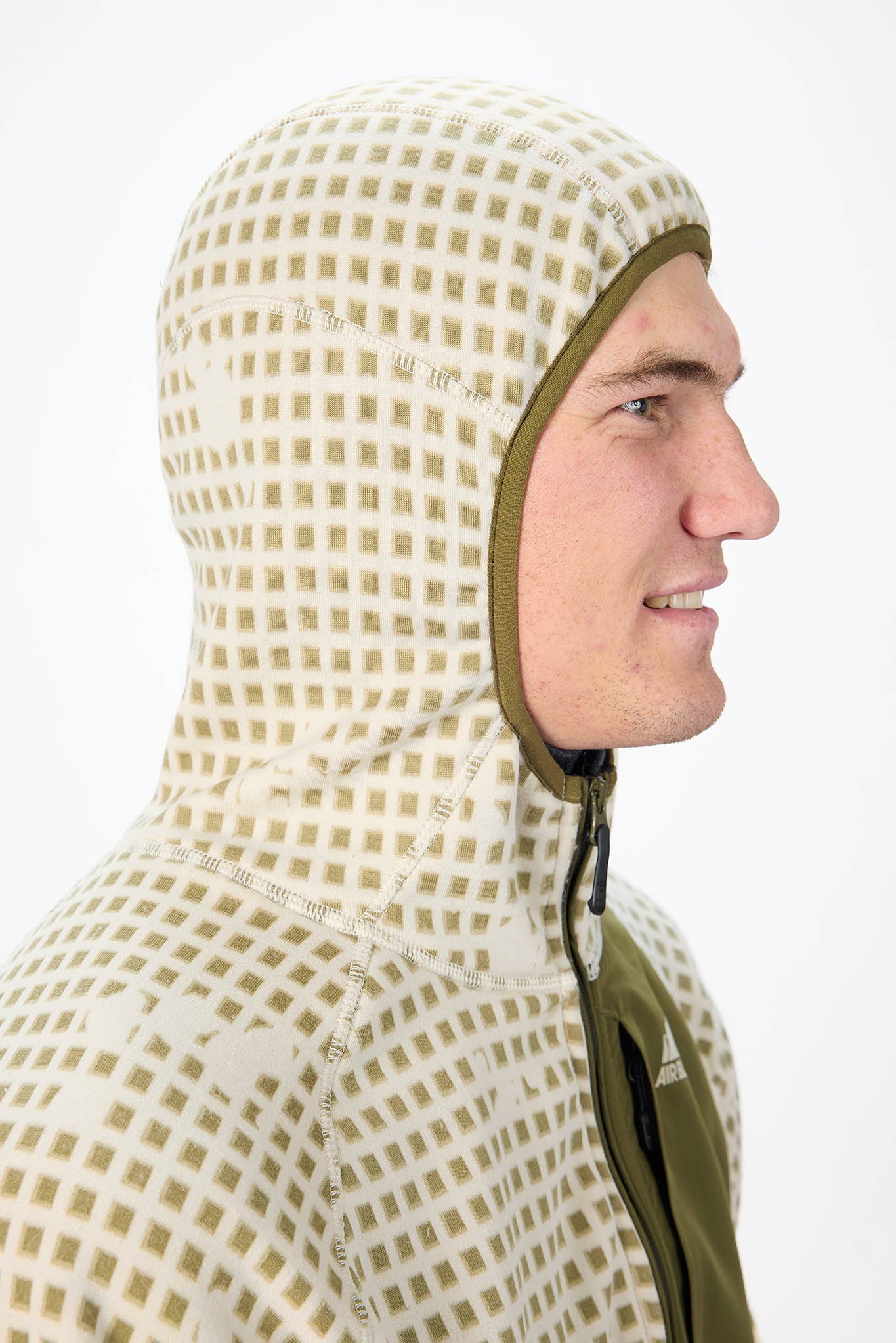 A man in profile wearing a hooded technical jacket with a beige and olive green checkered pattern and solid olive green trim.