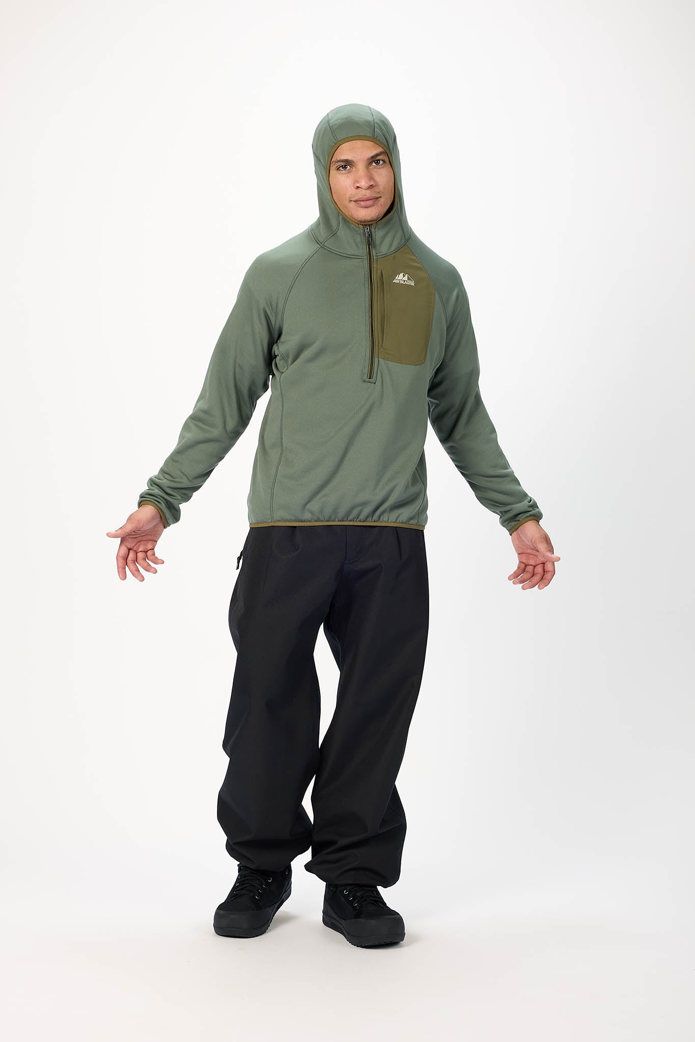 A man modeling an olive green hooded quarter-zip pullover jacket with a vertical chest pocket, paired with black pants and black shoes, against a plain white studio background.