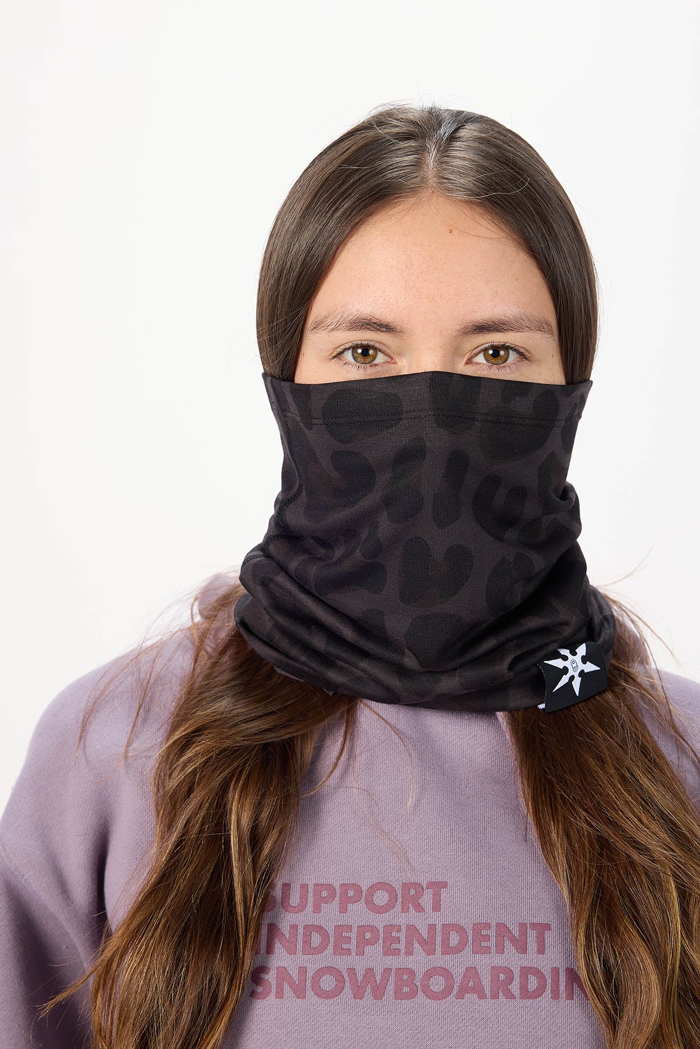 A woman wearing a black patterned neck gaiter mask with a small white snowflake logo, paired with a lavender sweatshirt against a plain white background.