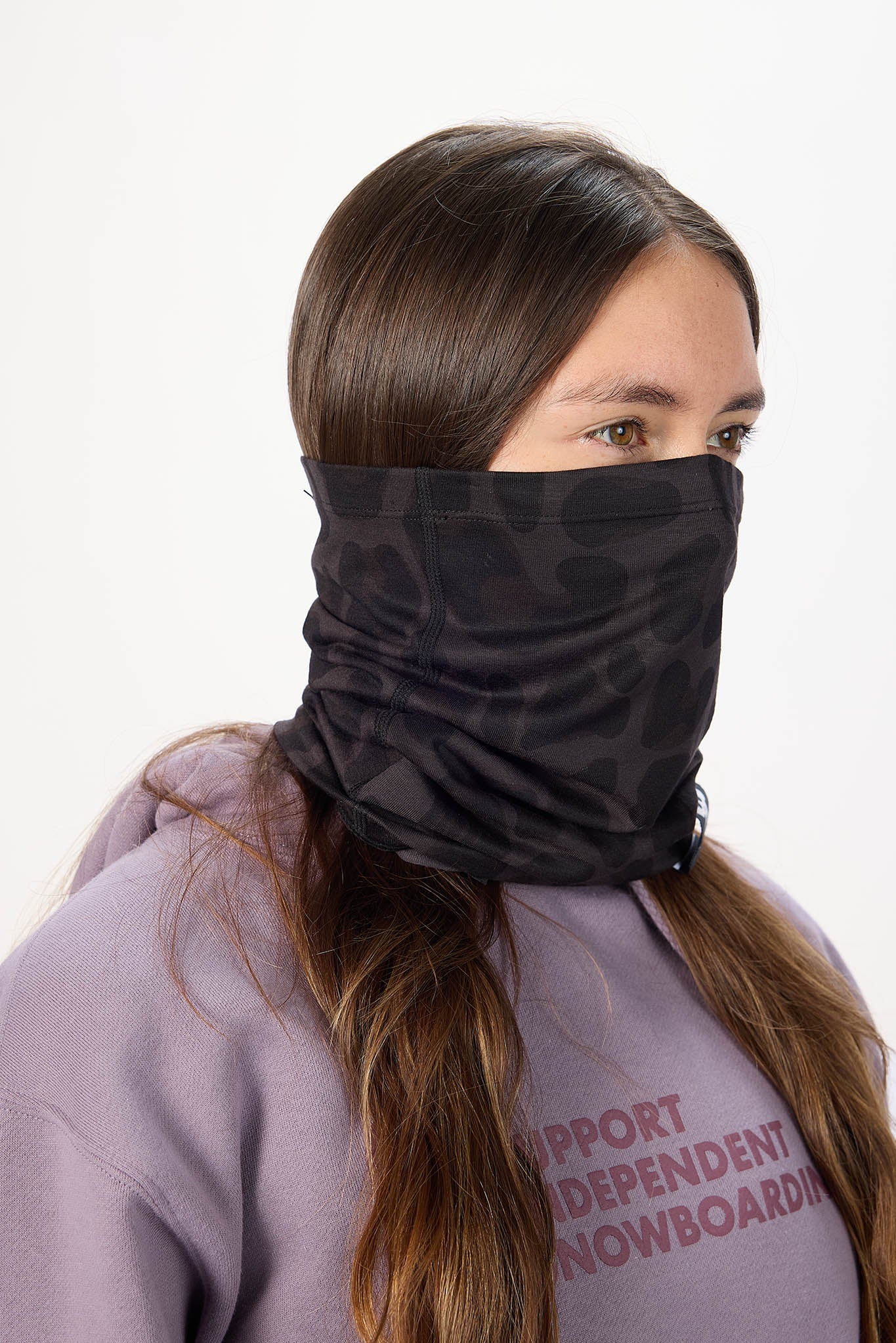 A woman wearing a black patterned neck gaiter or face covering pulled up over her nose and mouth, paired with a lavender hoodie against a plain white background.
