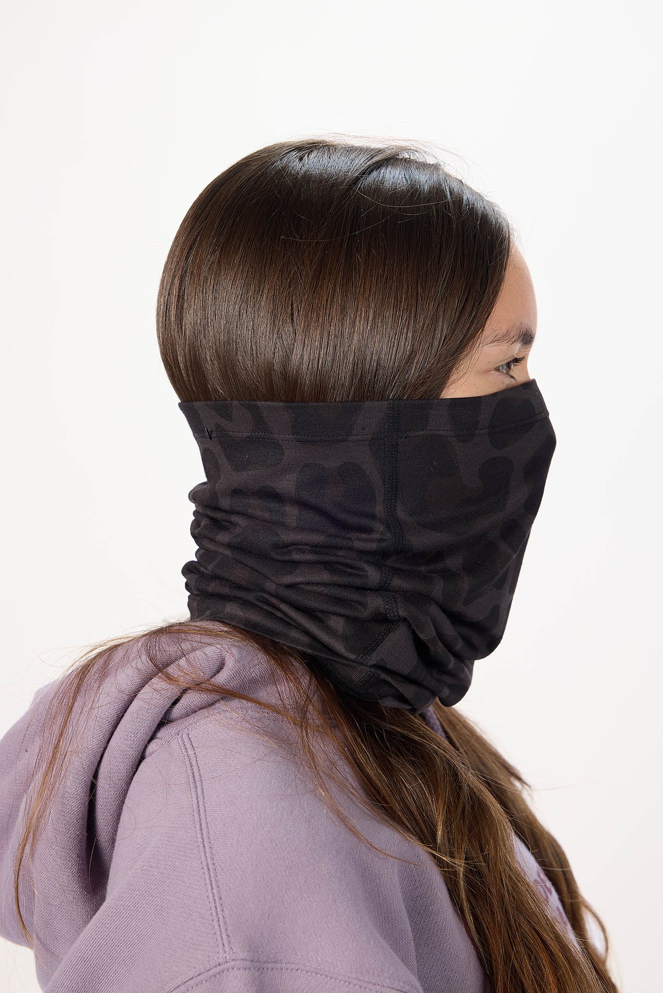 A person shown from the side wearing a dark camouflage-patterned neck gaiter or face mask over their nose and mouth, paired with a lavender hoodie against a plain white background.