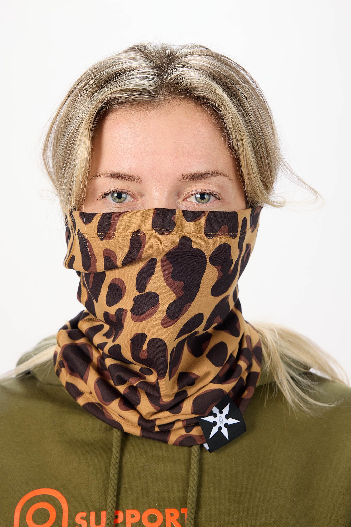A woman wearing a leopard-print neck gaiter over her nose and mouth, paired with an olive green hoodie. The neck gaiter features a tan and brown leopard pattern with a small black brand tag on the side.