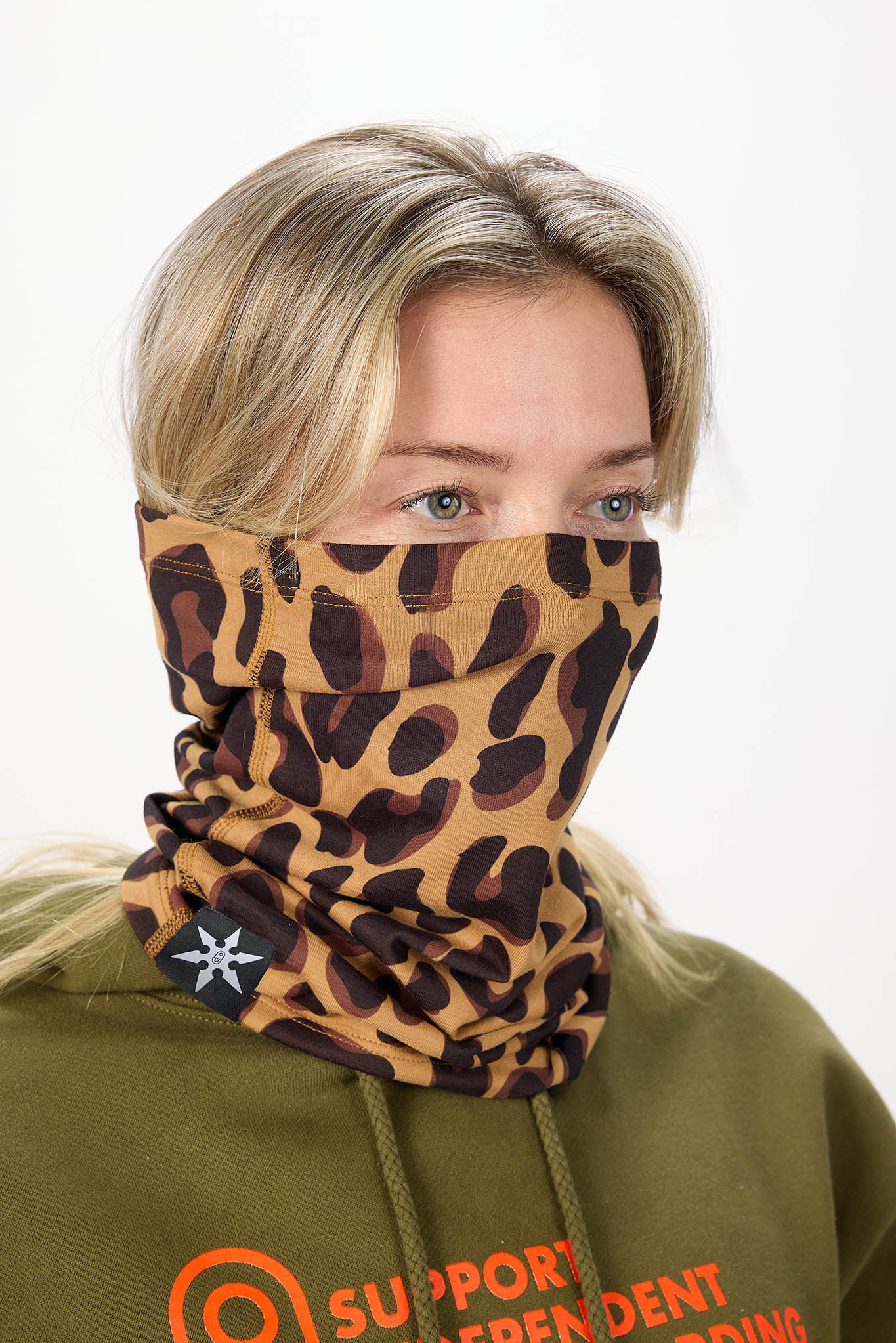 A woman wearing a leopard-print neck gaiter mask over her nose and mouth, paired with an olive green hoodie against a plain white background.