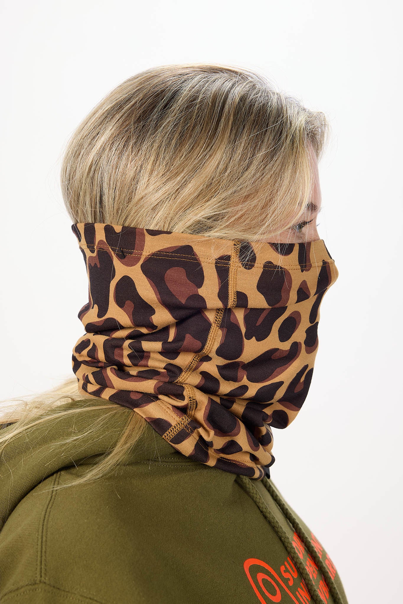 A side profile of a person wearing a leopard-print neck gaiter over their face and a green hoodie against a plain white background.