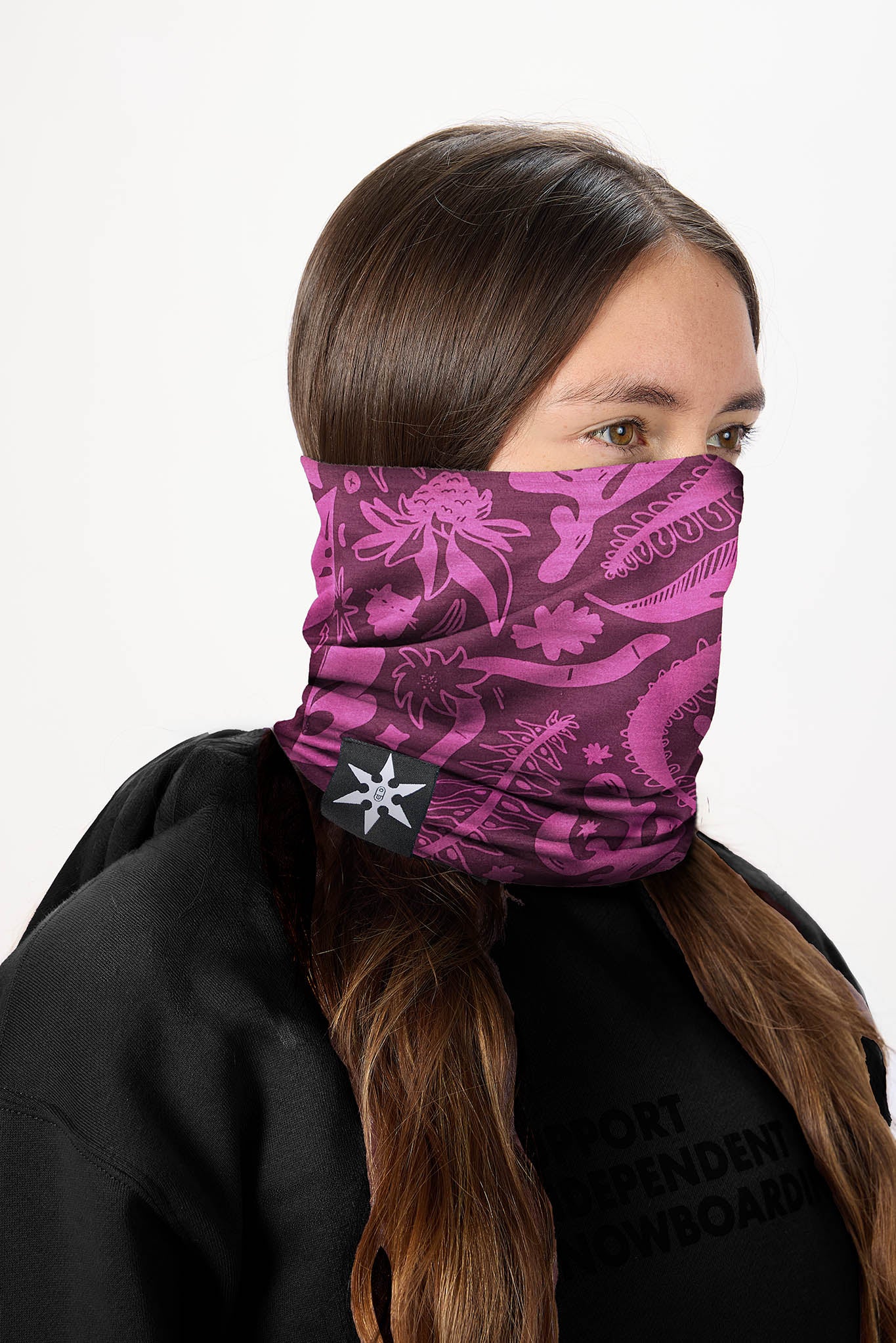 A woman wearing a pink and purple patterned neck gaiter mask with a white star logo, paired with a black hoodie against a plain white background.