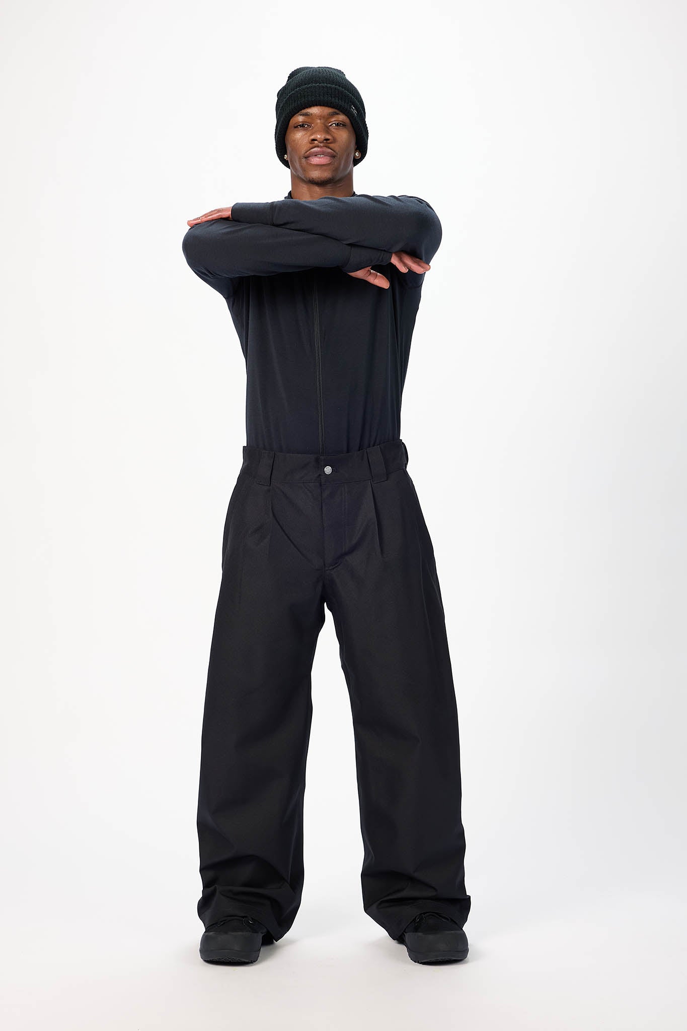 A model wearing a black beanie, black long-sleeve top, and black wide-leg pants stands with arms crossed against a plain white studio background.