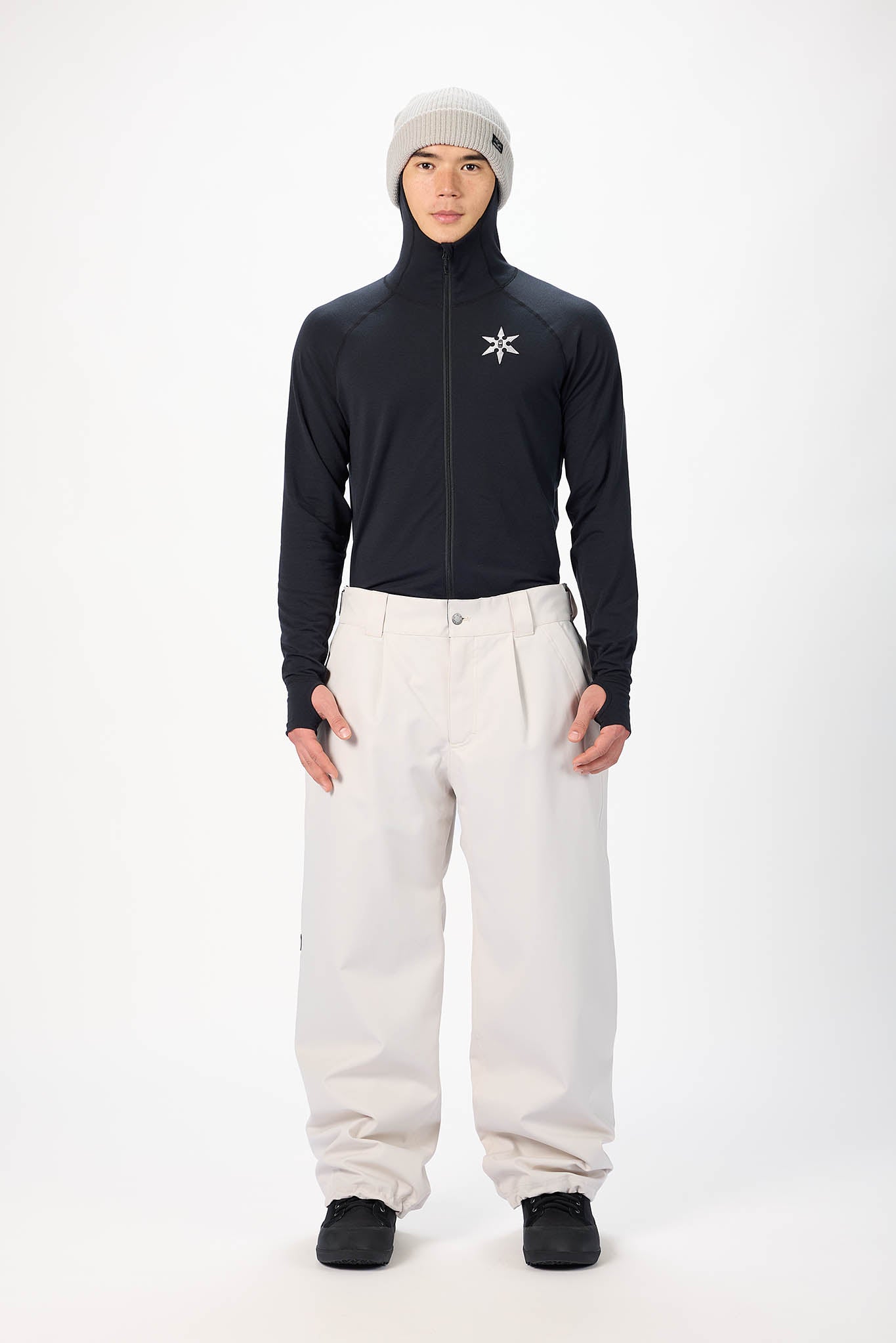 A model stands against a white studio background wearing a black long-sleeve zip-up athletic top with a white star logo on the chest, off-white snow pants, black shoes, and a light gray knit beanie.