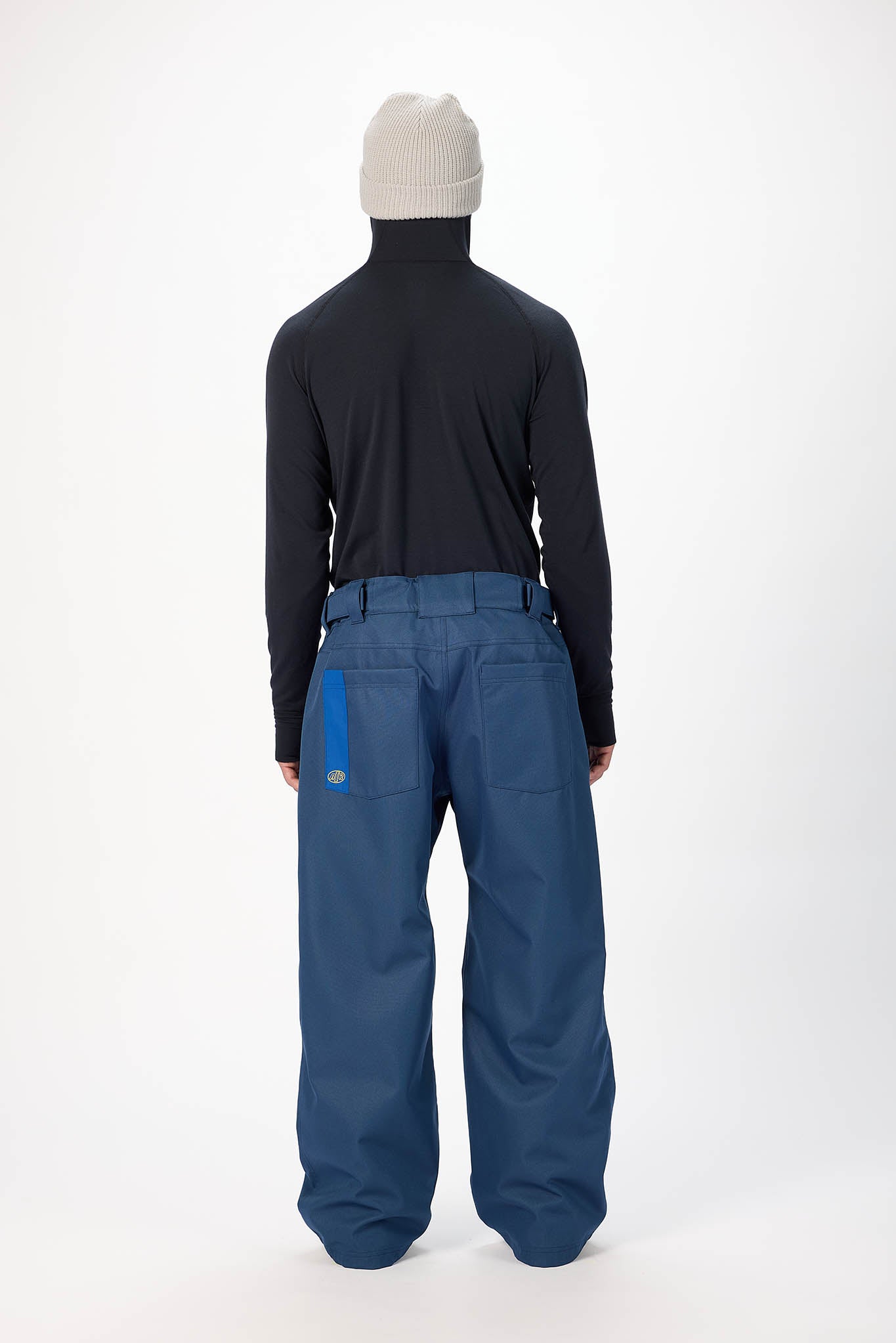 A model shown from the back wearing a black turtleneck, a light beige knit beanie, and wide-leg blue snow pants with a bright blue vertical accent on the back pocket.