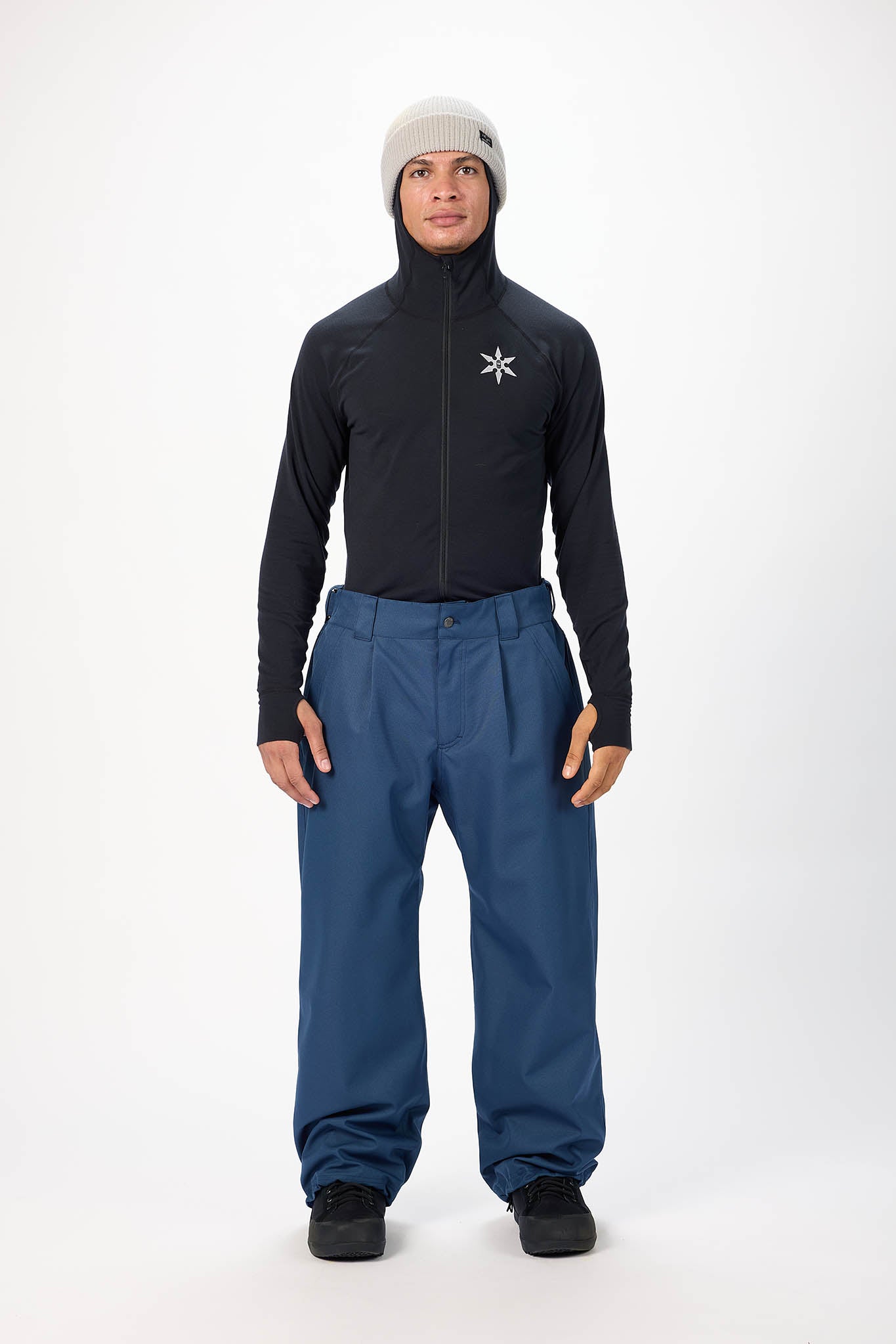 A man modeling a black long-sleeve technical jacket with a white logo on the chest, paired with blue snow pants, a light gray beanie, and black boots, against a plain white studio background.