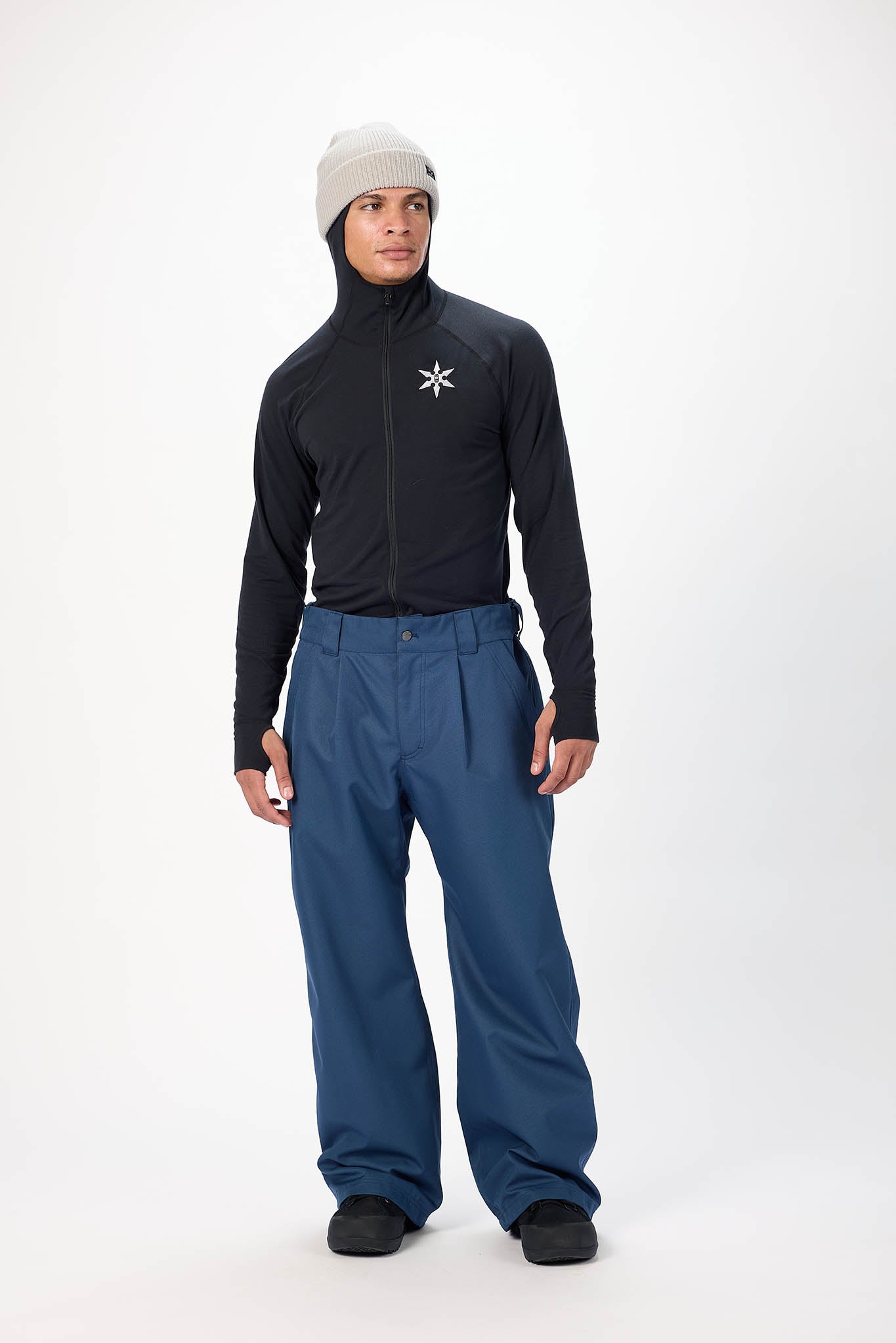 A man modeling a black long-sleeve technical jacket with a white logo on the chest, paired with blue snow pants, black shoes, and a light gray beanie, against a plain white studio background.