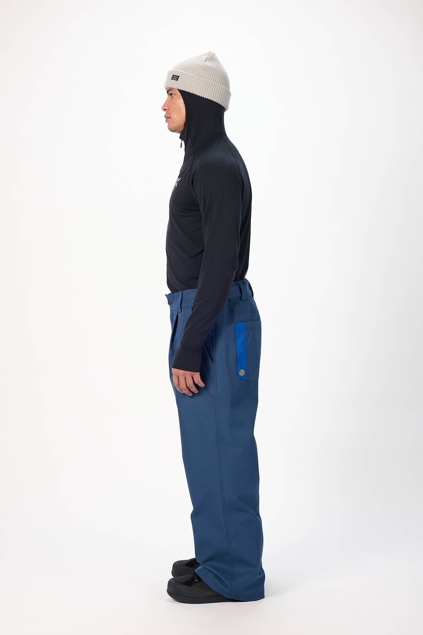 A side-profile studio shot of a person wearing a light gray beanie, a dark navy long-sleeve top, and blue snow pants with bright blue accents, standing against a plain white background.