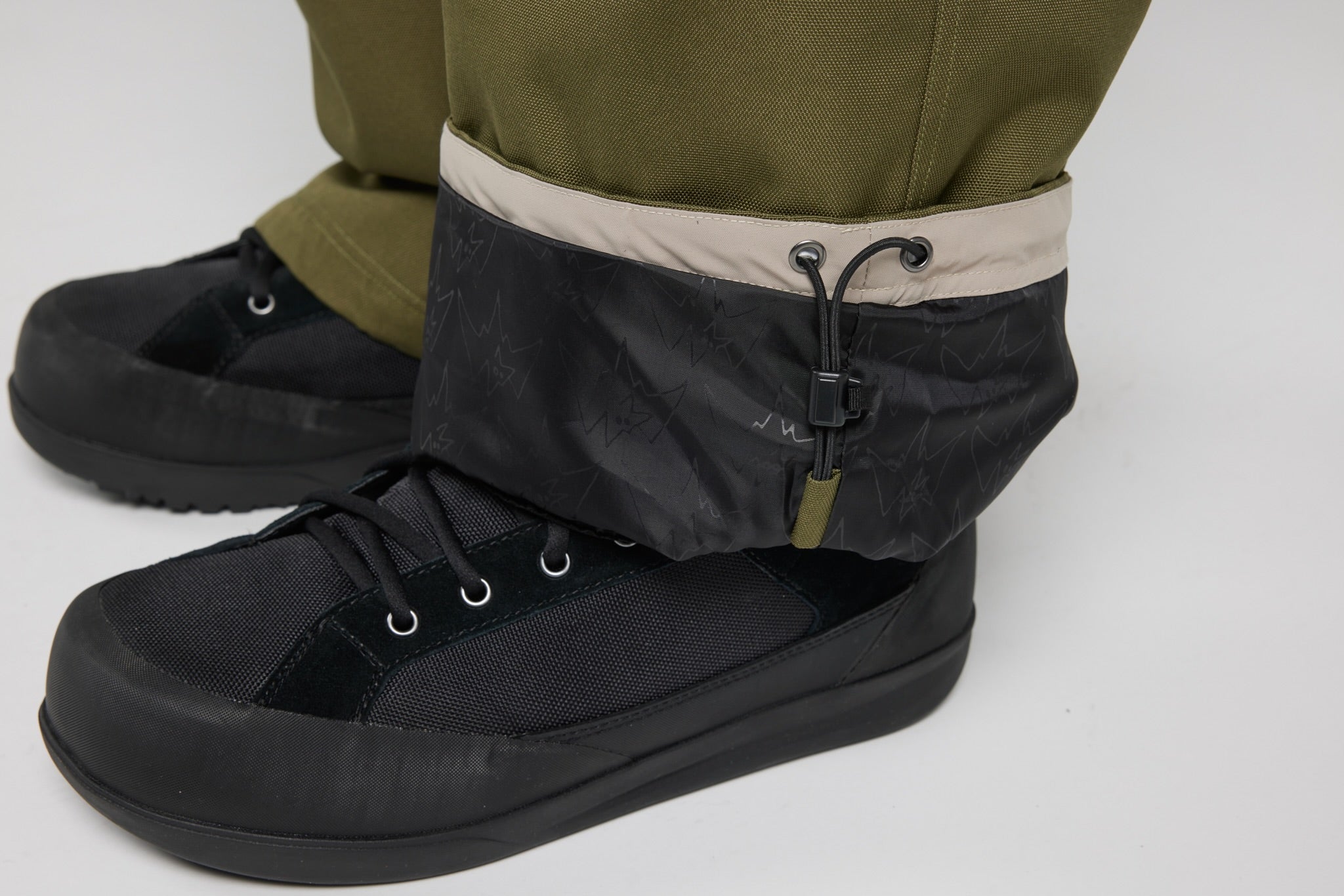 Black low-top lace-up boots with a dark patterned cuff and a beige inner lining, shown worn with olive green pants on a light gray studio background.