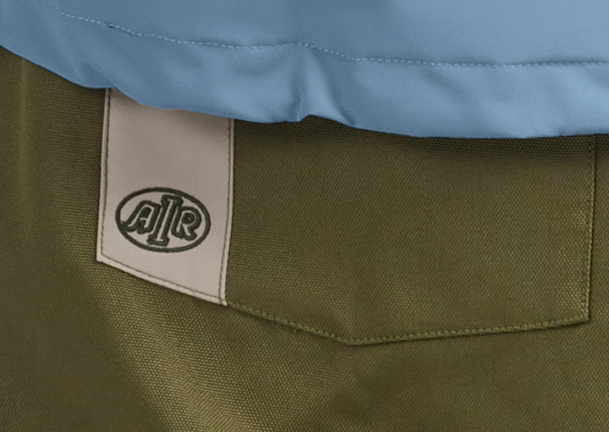 Close-up of a garment showing an olive green fabric section with a beige brand patch reading AJR, plus a light blue section at the top.