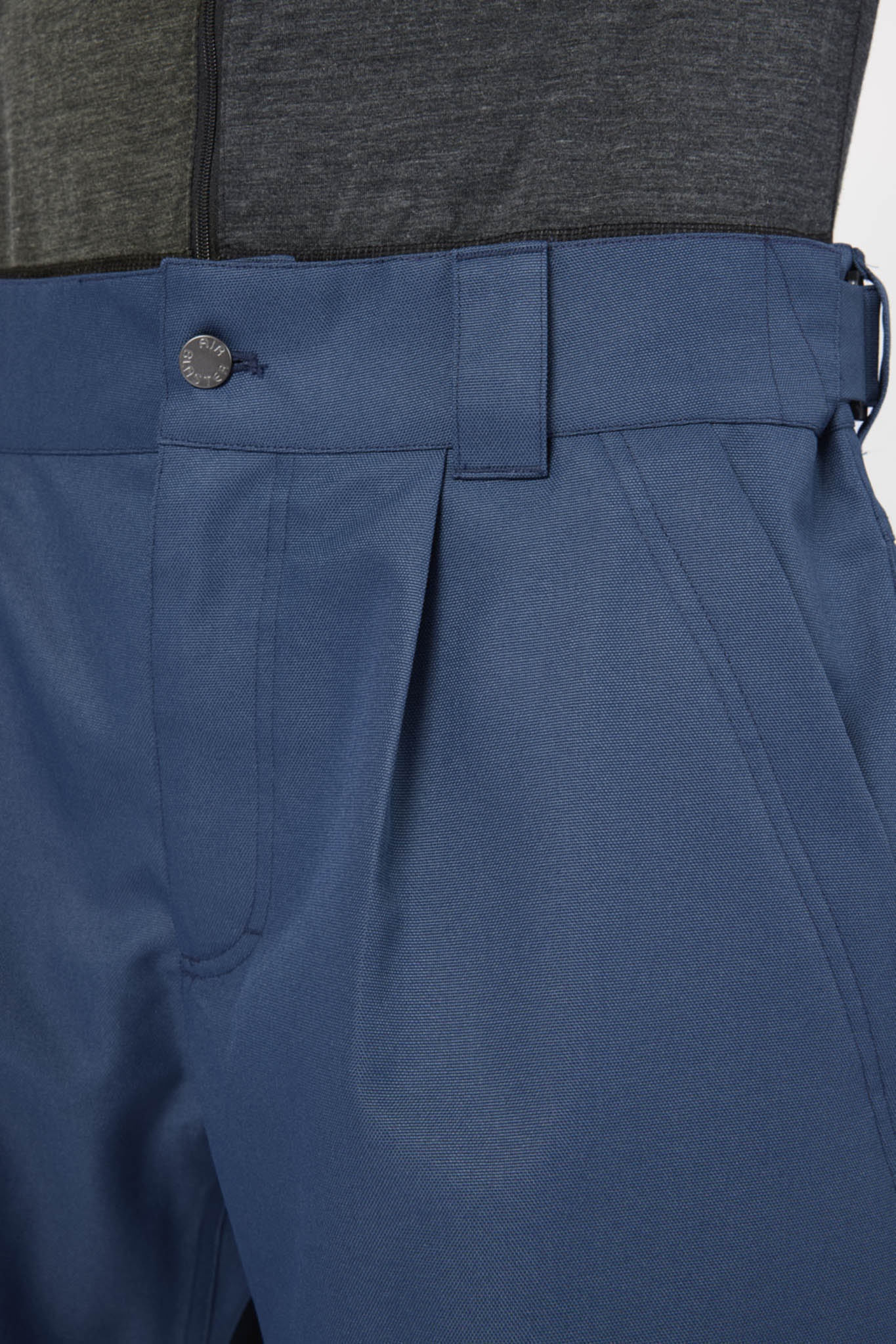 Close-up of blue tailored trousers showing the waistband, belt loop, button, front seam, and side pocket, with a dark gray top partially visible at the top edge.