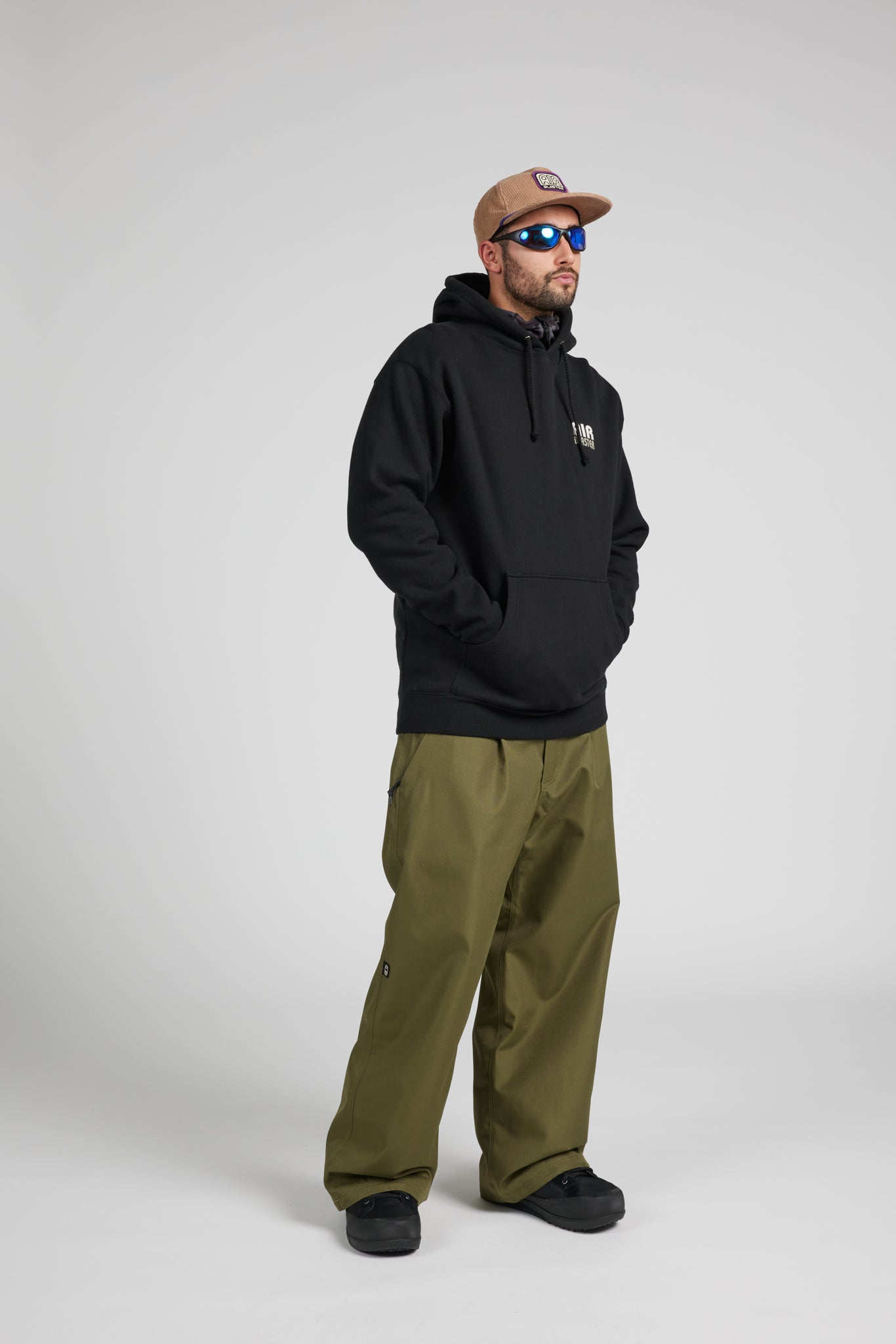 A man wearing a black pullover hoodie, olive green pants, a brown baseball cap, and blue-tinted sunglasses, standing against a light gray studio background.