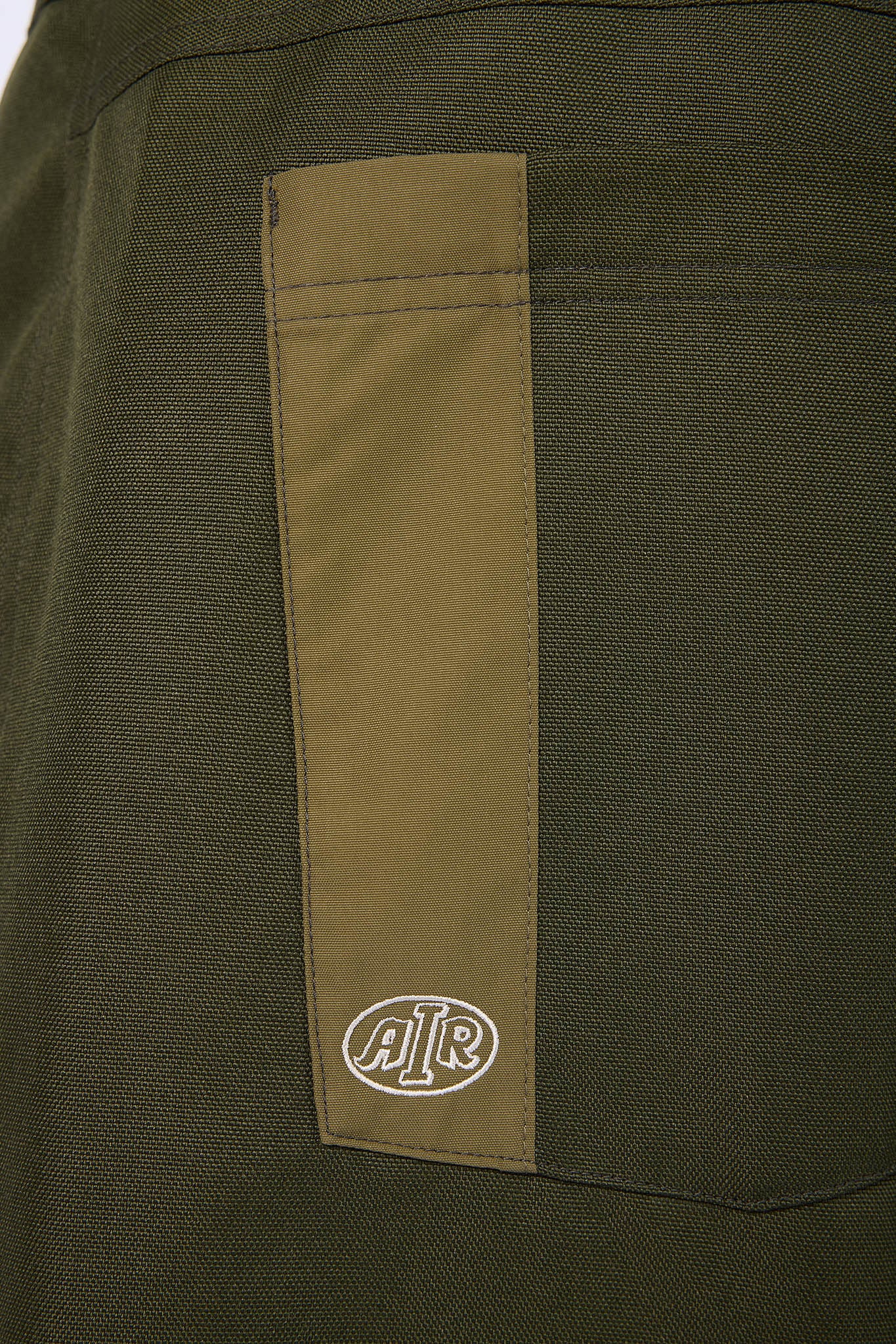 Close-up of a dark olive green fabric garment featuring a vertical tan patch pocket with a white embroidered circular AIR logo.