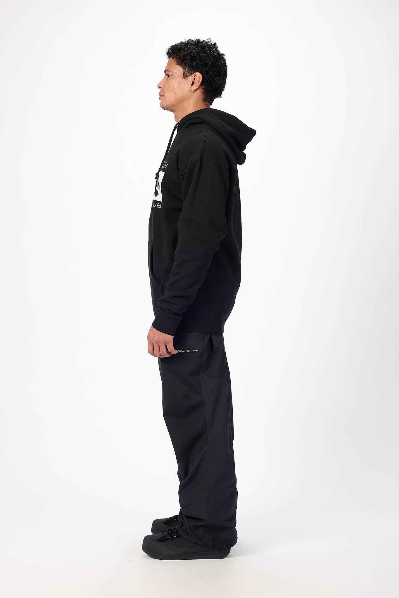 A man in a black hoodie and black pants stands in profile against a plain white studio background.