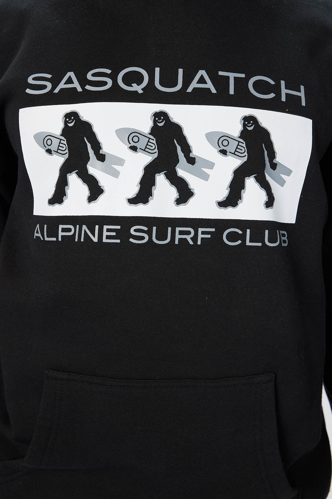 Close-up of a black pullover hoodie featuring a large front graphic with the text 'SASQUATCH' and 'ALPINE SURF CLUB' in white and gray, plus three silhouettes of a bigfoot-like figure carrying a surfboard inside a white rectangular panel.