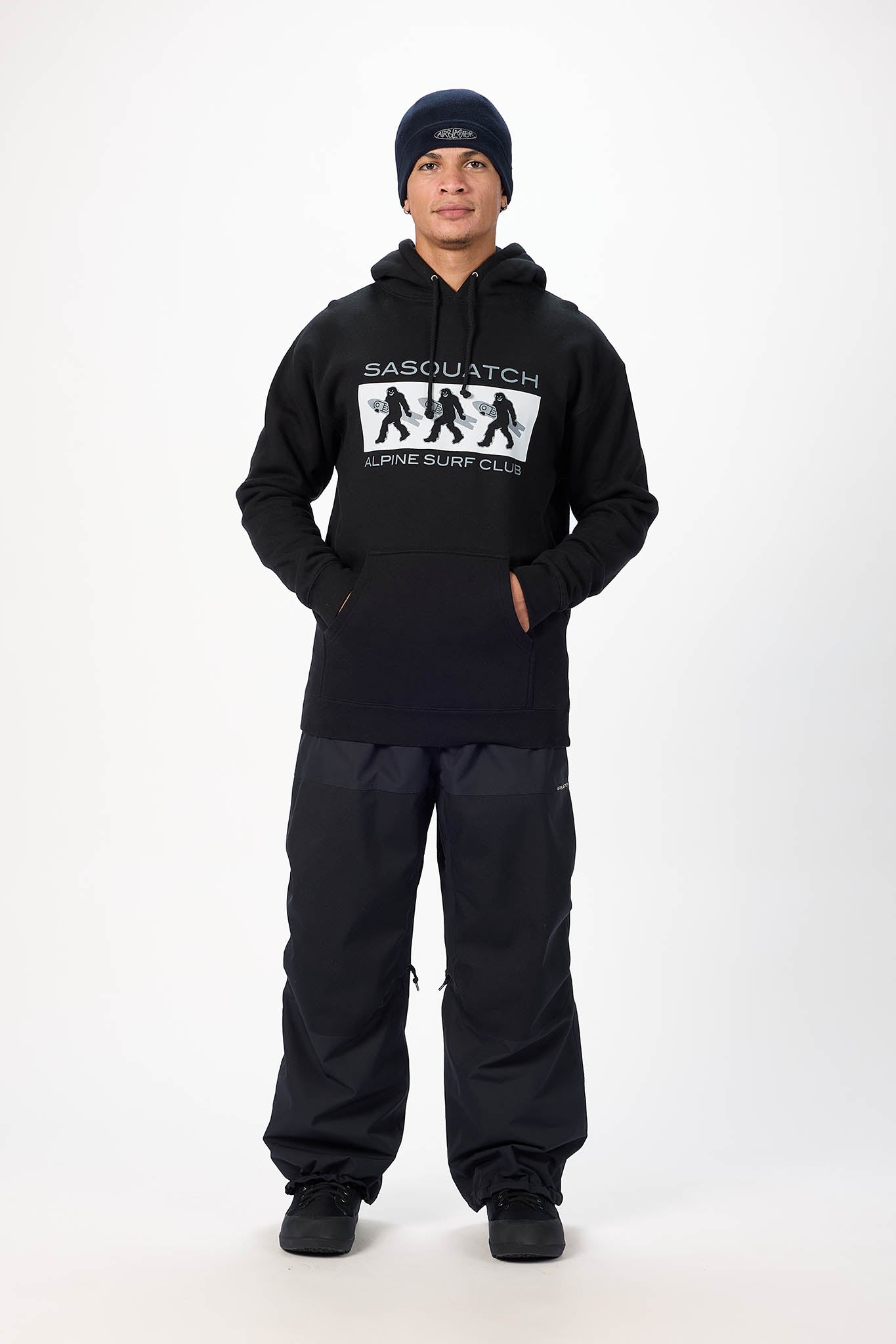 A man modeling a black Sausatch Alpine Surf Club hoodie and matching black pants against a plain white studio background.