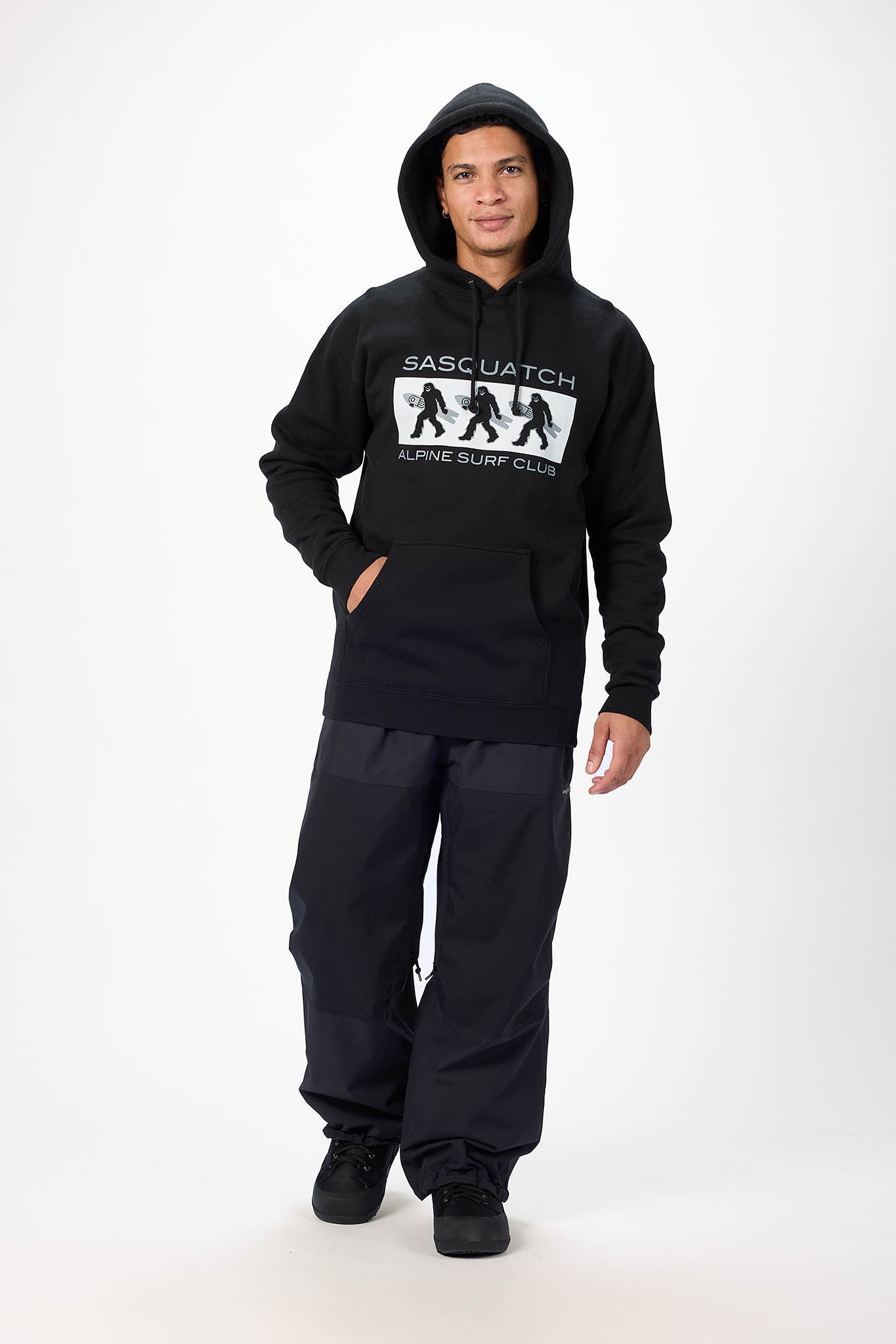 A man modeling a black pullover hoodie with a white graphic print on the chest that reads 'SASC. WATCH ALIVE SURF CLUB' with four silhouettes, paired with black pants and black shoes, against a plain white studio background.