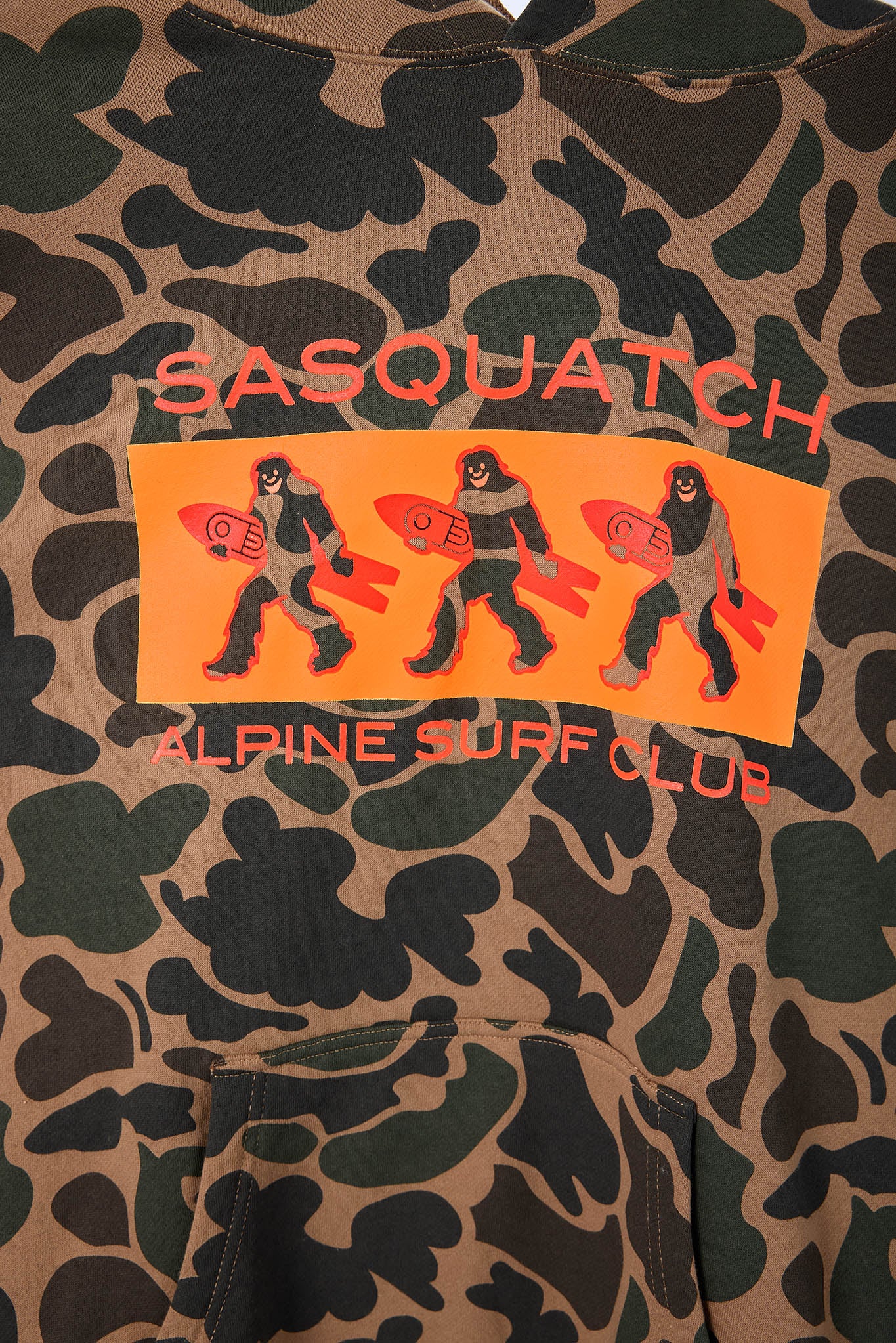 Close-up of a camouflage sweatshirt featuring a central graphic with three cartoon figures carrying orange surfboards and the text 'SASQUATCH' and 'ALPINE SURF CLUB'.