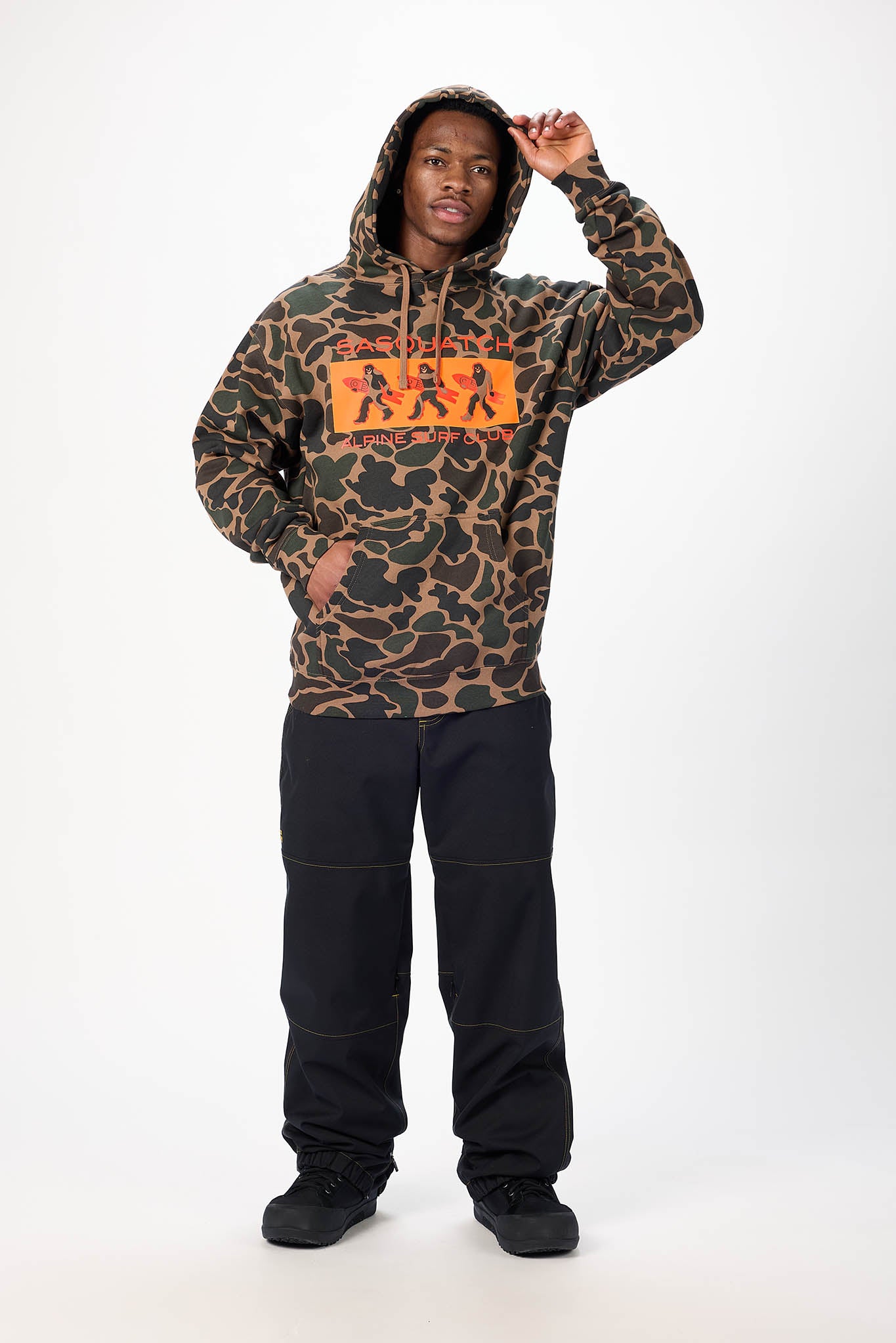 A man modeling a camouflage hoodie with a prominent orange graphic on the chest, paired with black pants and black sneakers, against a plain white studio background.