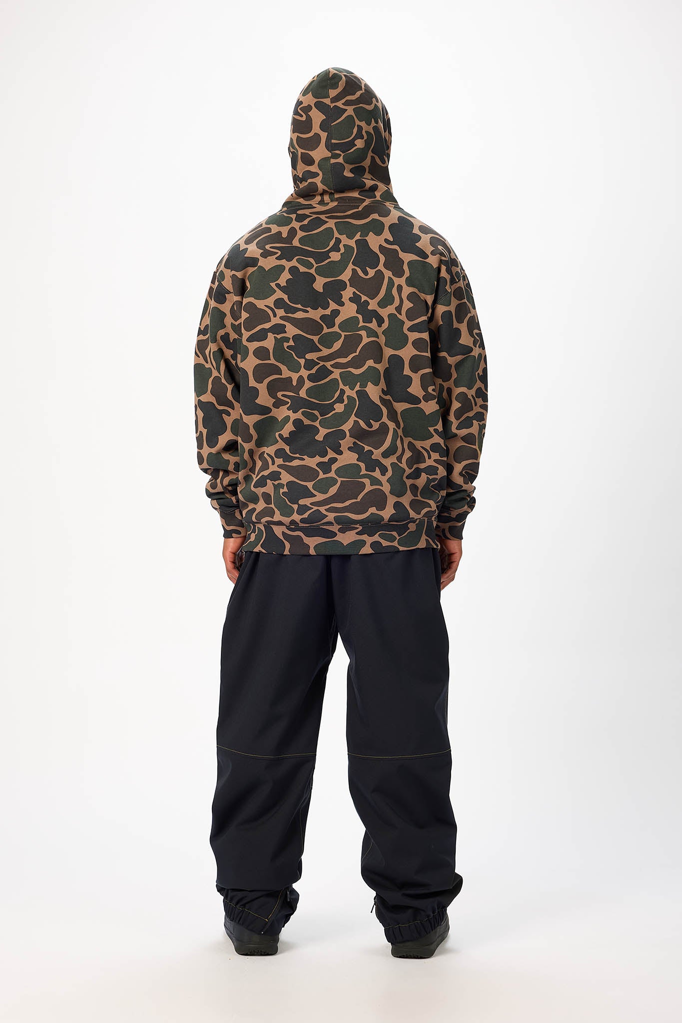A person shown from the back wearing a camouflage-patterned hooded jacket and black pants against a plain white studio background.