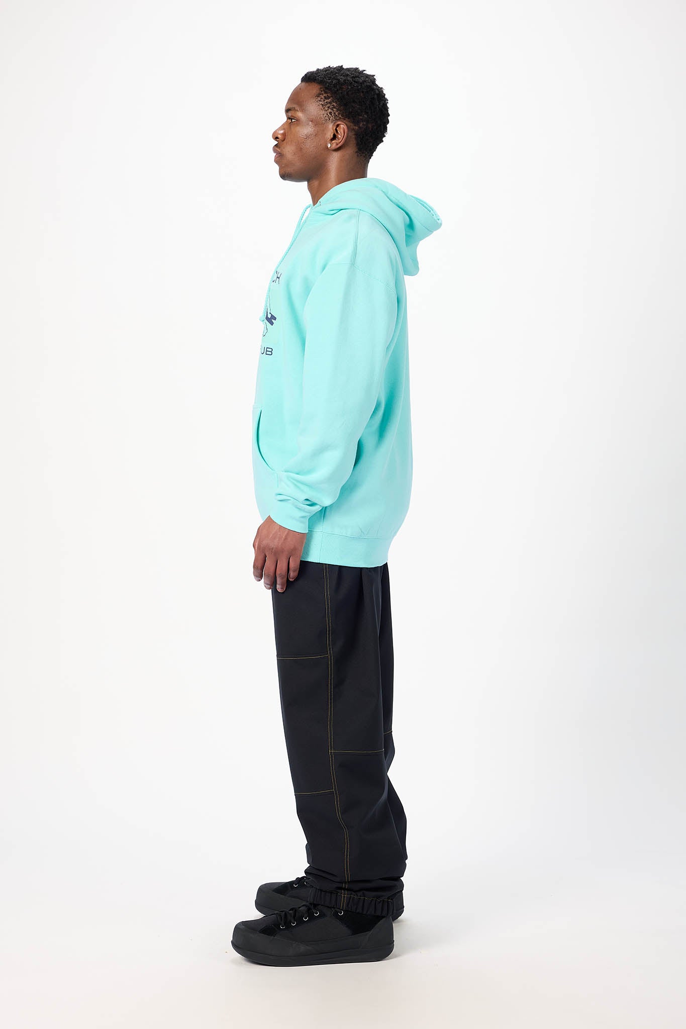 A man in profile wearing a light aqua hooded sweatshirt, black pants, and black sneakers, standing against a plain white studio background.