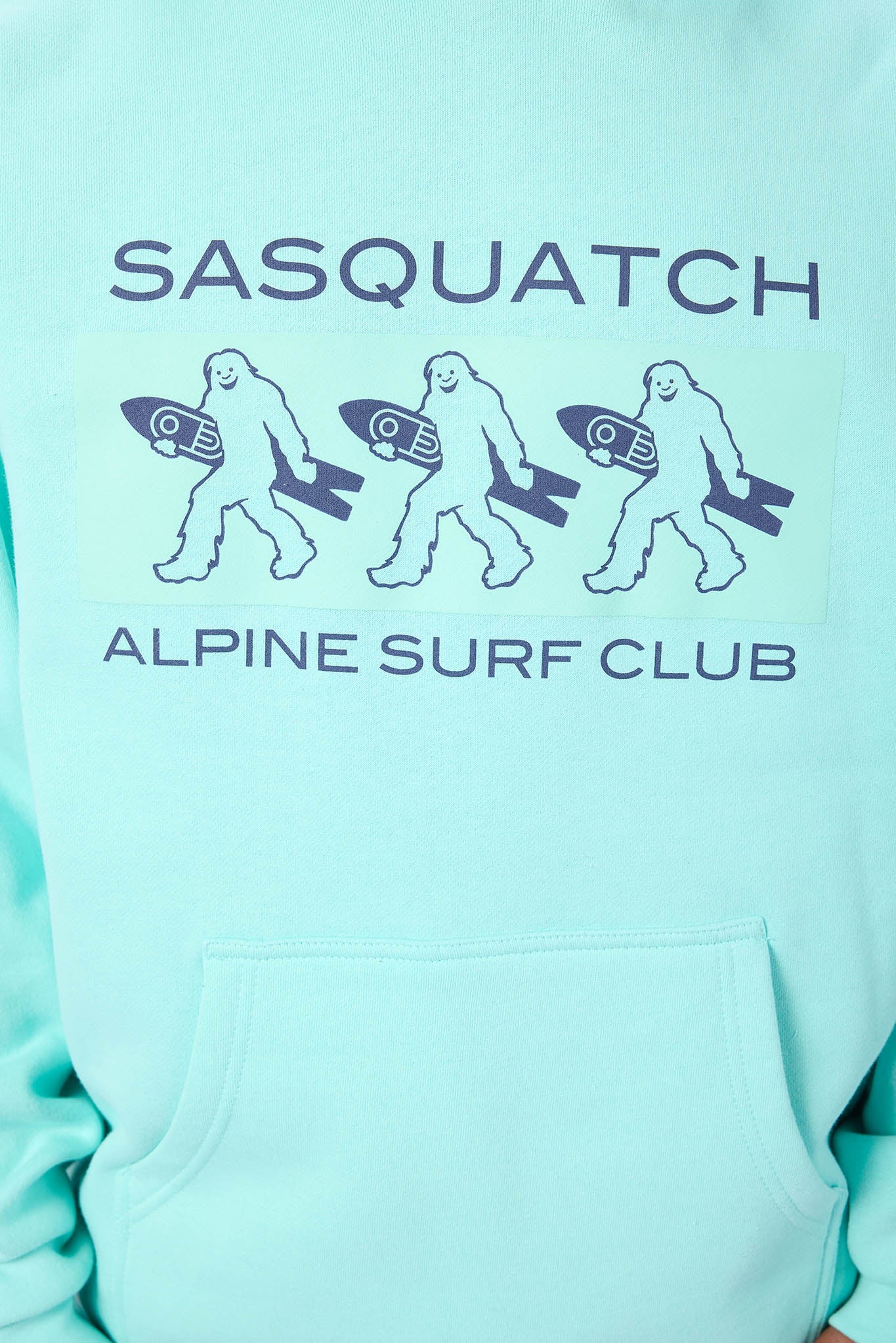 A close-up of a light aqua hoodie featuring a graphic print of three walking sasquatch figures carrying surfboards, with the text 'SASQUATCH' above and 'ALPINE SURF CLUB' below.