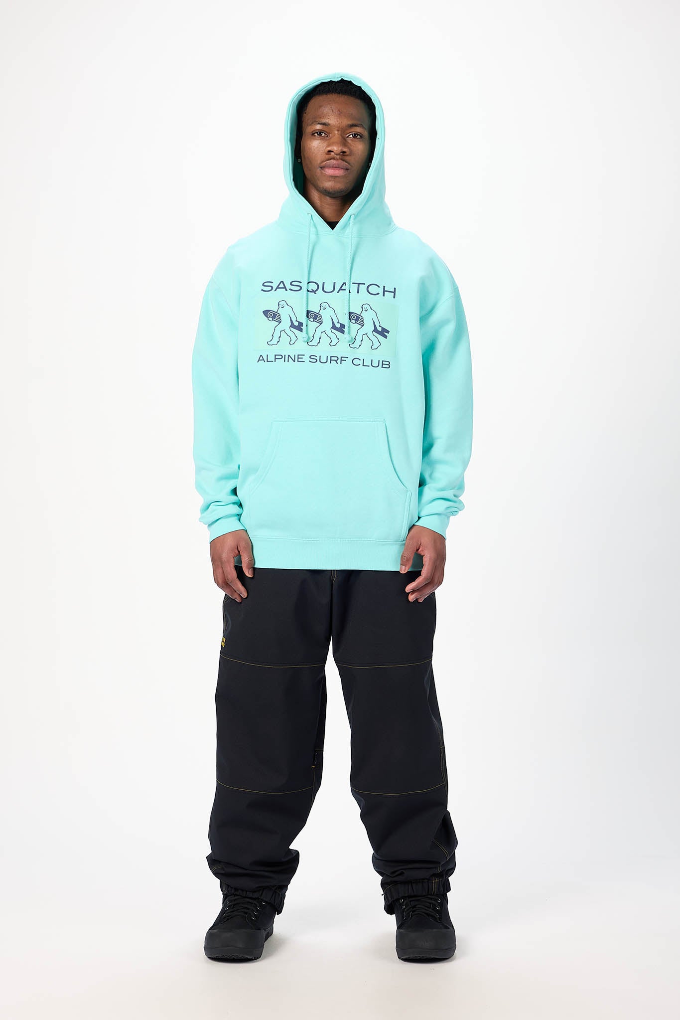 A model wearing a light aqua blue hooded sweatshirt with a black graphic print on the chest reading 'SASQUATCH ALPINE SURF CLUB', paired with black sweatpants and black sneakers, against a plain white studio background.