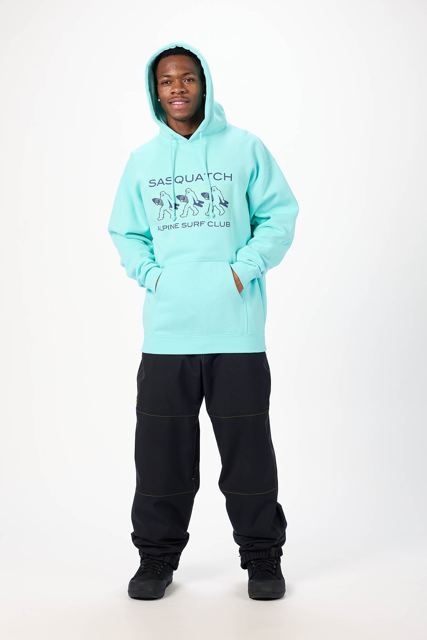 A model wearing a light aqua/turquoise pullover hoodie with 'SASQUATCH SURF CLUB' printed on the chest, paired with black sweatpants and black sneakers, standing against a plain white studio background.