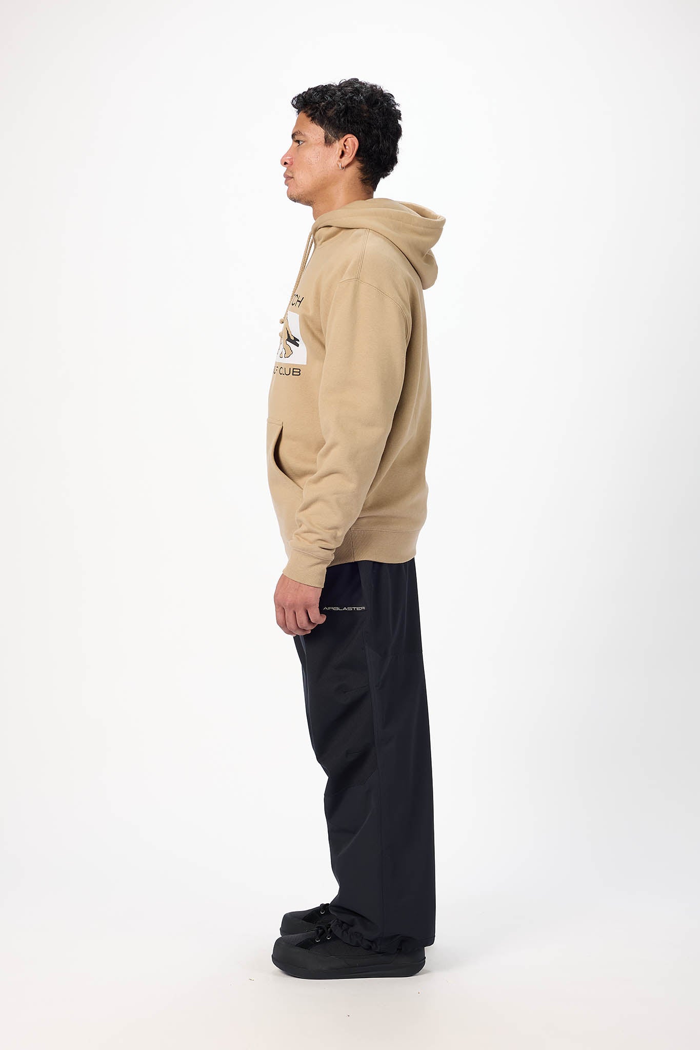 A side-profile studio shot of a young man wearing a beige hoodie with a graphic on the chest and dark navy wide-leg sweatpants, standing against a plain white background.