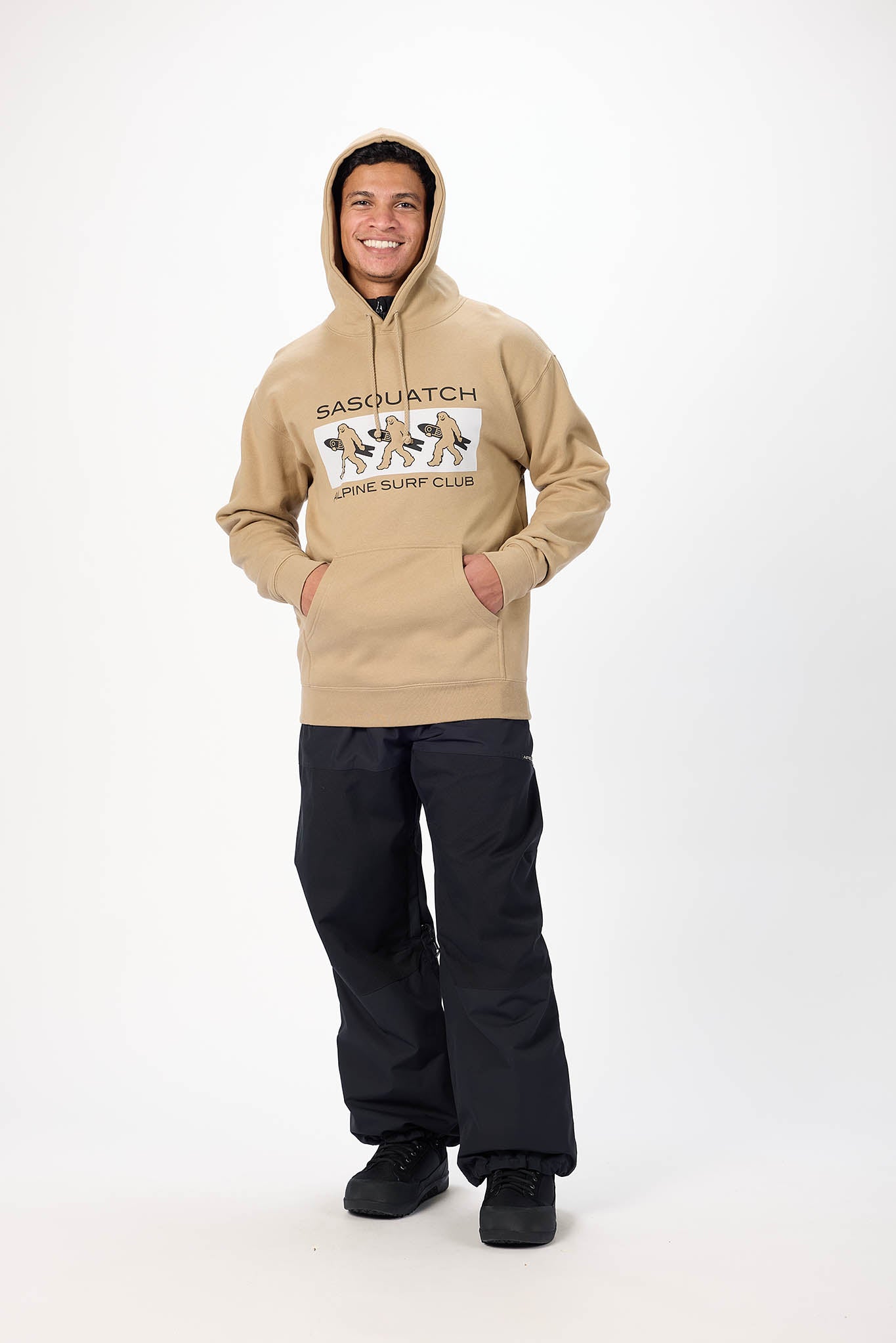 A man wearing a beige hooded sweatshirt with a 'SASQUATCH LITTLE SURF CLUB' graphic on the chest, paired with black snow pants and black shoes, standing against a white studio background.