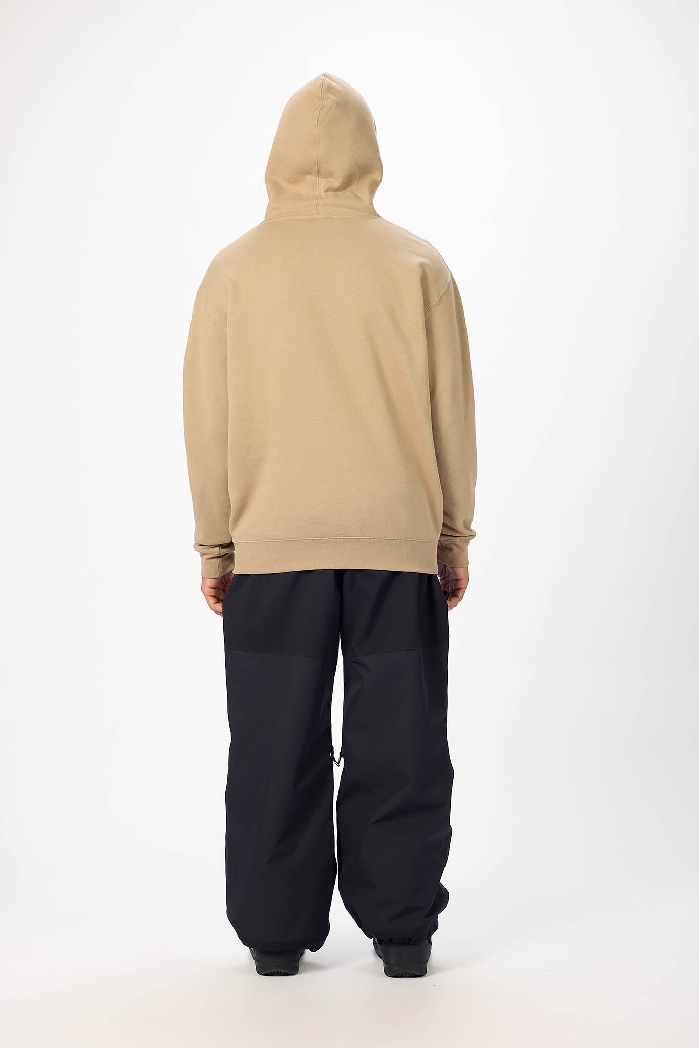 A person shown from the back wearing a beige hooded sweatshirt and loose black pants, standing against a plain light gray studio background.