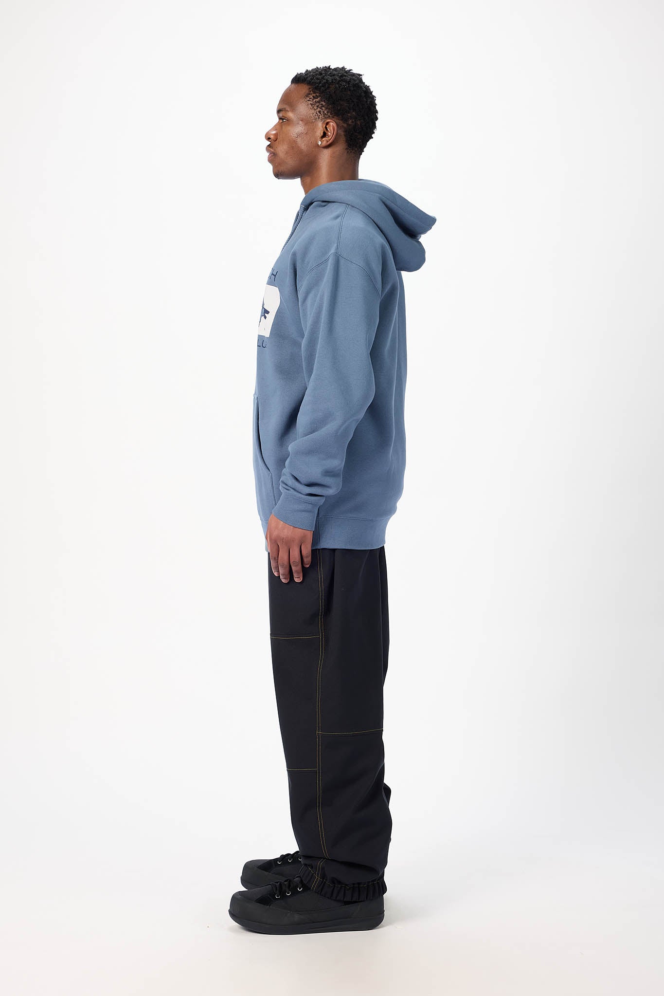 A man in profile wearing a dusty blue pullover hoodie with a white chest patch, black pants, and black sneakers, standing against a plain white studio background.