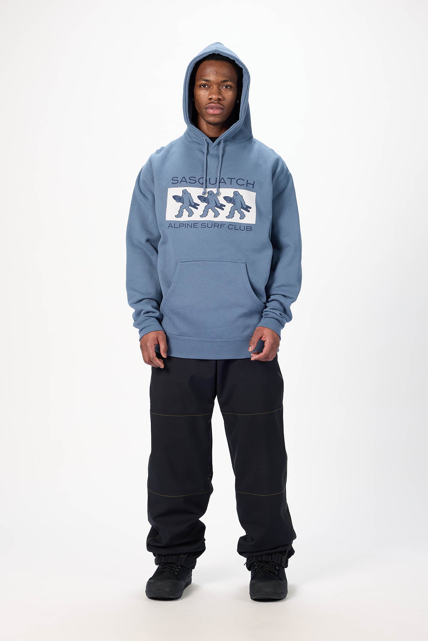 A model stands against a white studio background wearing a light blue hooded sweatshirt with a white and blue graphic on the chest that reads 'SASQUATCH' and 'MORPHING SHIP CROWN', paired with black sweatpants and black sneakers.