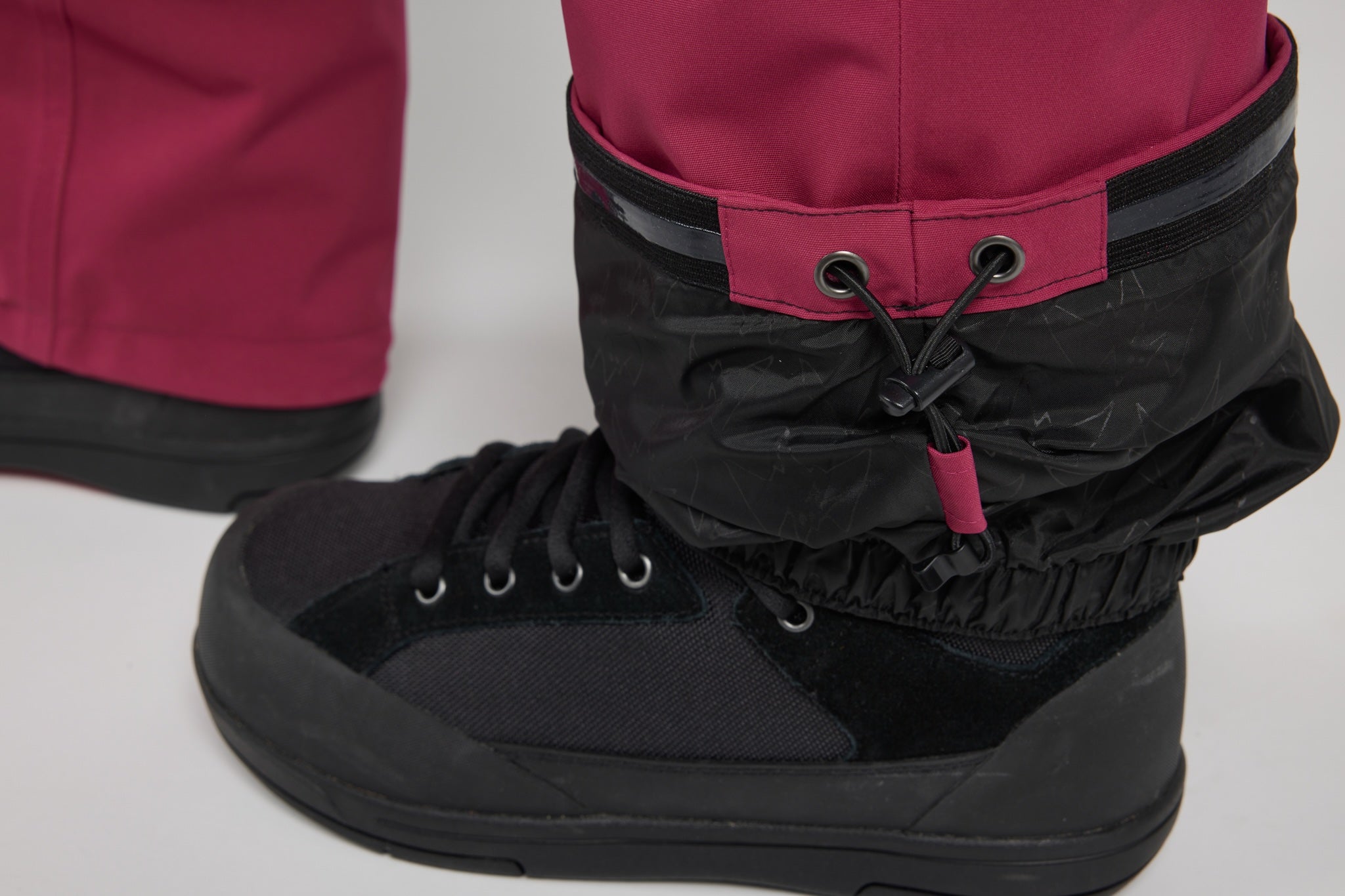 A close-up of a black lace-up boot with a black gaiter cuff and a small magenta pull tab, paired with matching magenta pants on a light gray studio background.